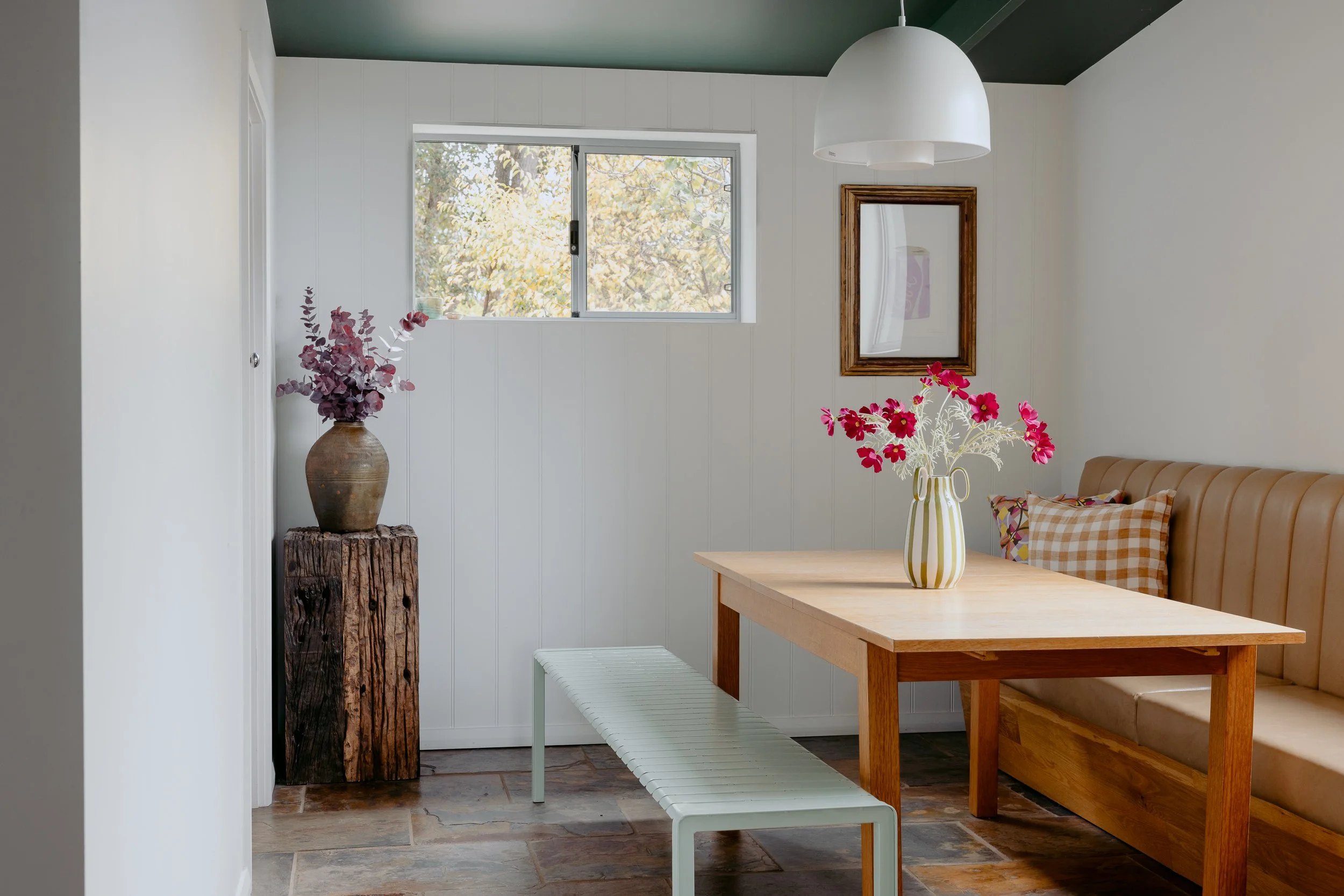 Interior of a home with a white wall, green ceiling, and a window showing trees outside. There is a tall vase with purple flowers on a wooden stump, a striped vase with pink flowers on a wooden table, and a mirror hanging on the wall. Part of a tan sofa and colorful cushions are also visible.