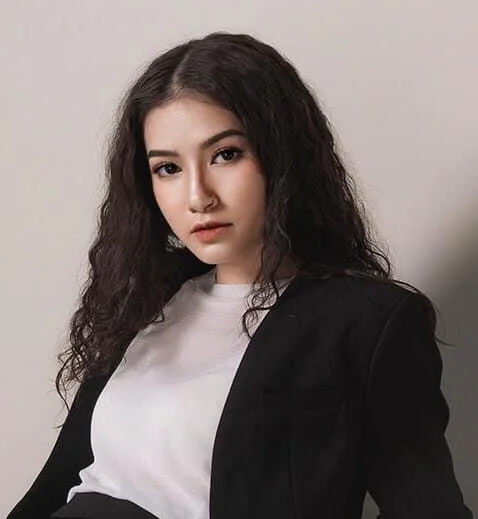 A young woman with long, curly dark hair wearing a white shirt and black blazer against a plain background.