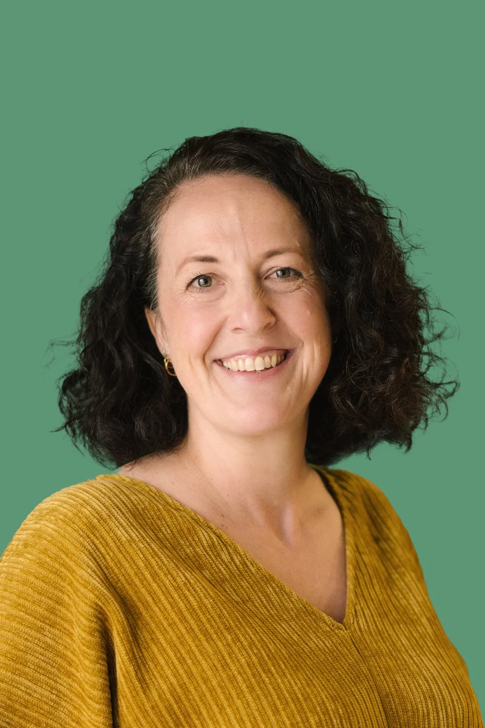 Portrait of a woman with curly dark hair wearing a mustard yellow top, smiling, against a solid green background.