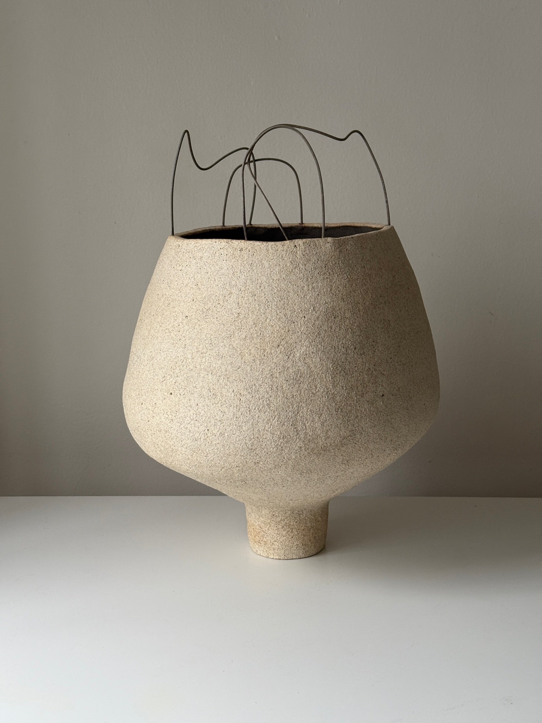 A textured beige ceramic vase with a narrow base and large rounded body, containing thin, wiry metal decor at the top.