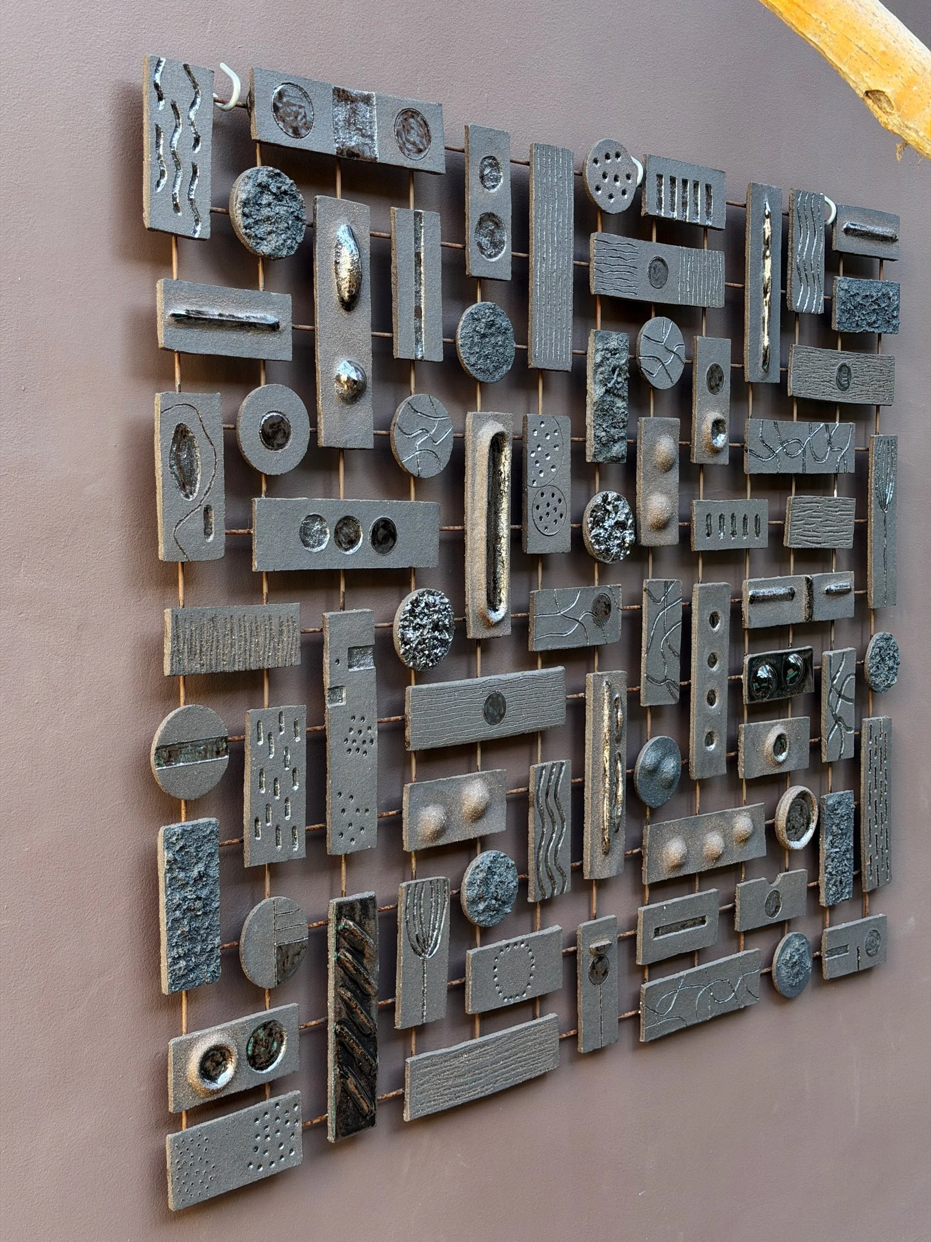 Ceramic tile wall installation by Anna Shipulina, Study I — hand-built charcoal-toned tiles with carved textures and metallic accents, mounted on a metal grid.