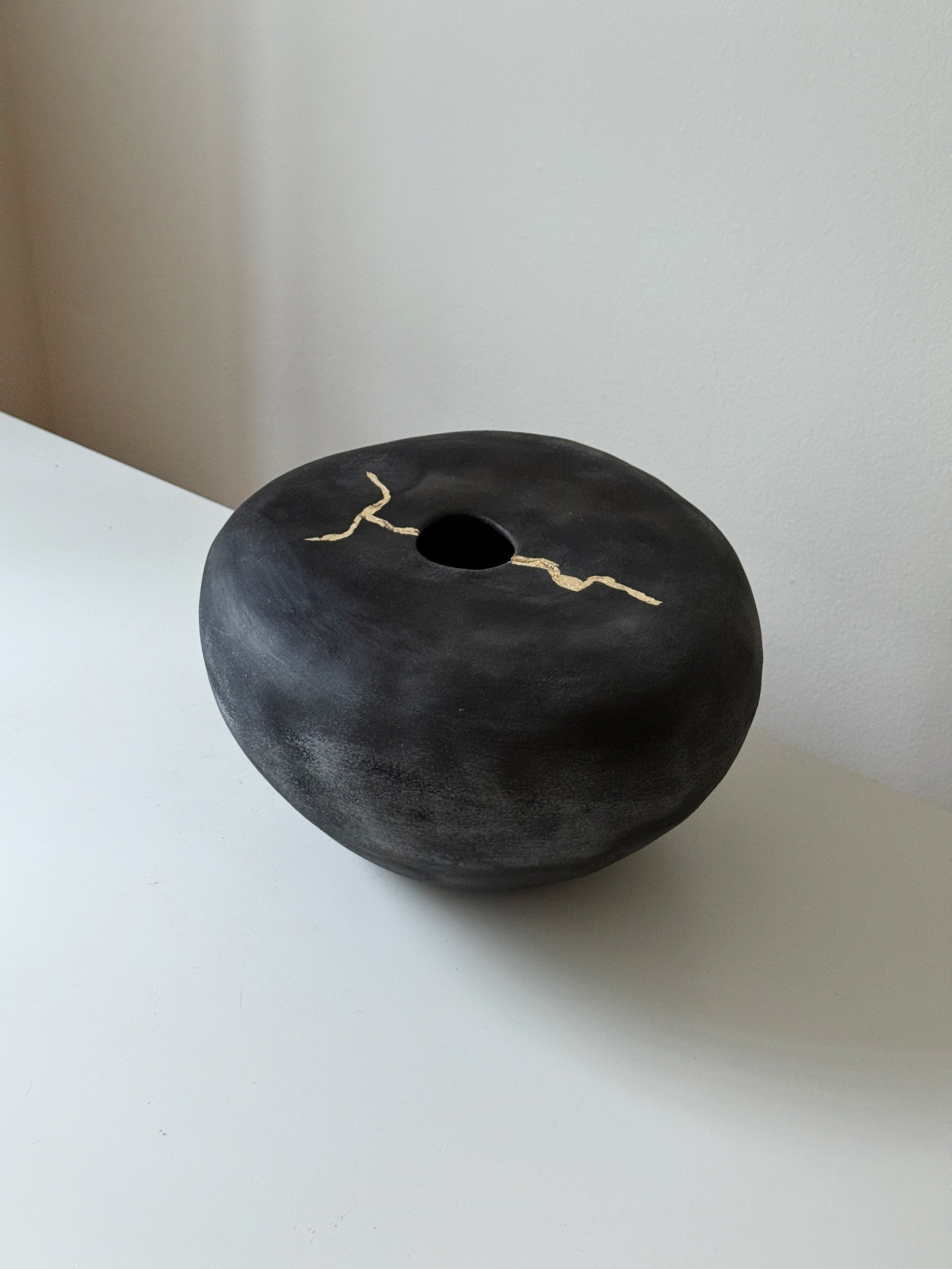 Rounded matte black ceramic vase with a central opening and a gold seam running across the top, on a white surface.