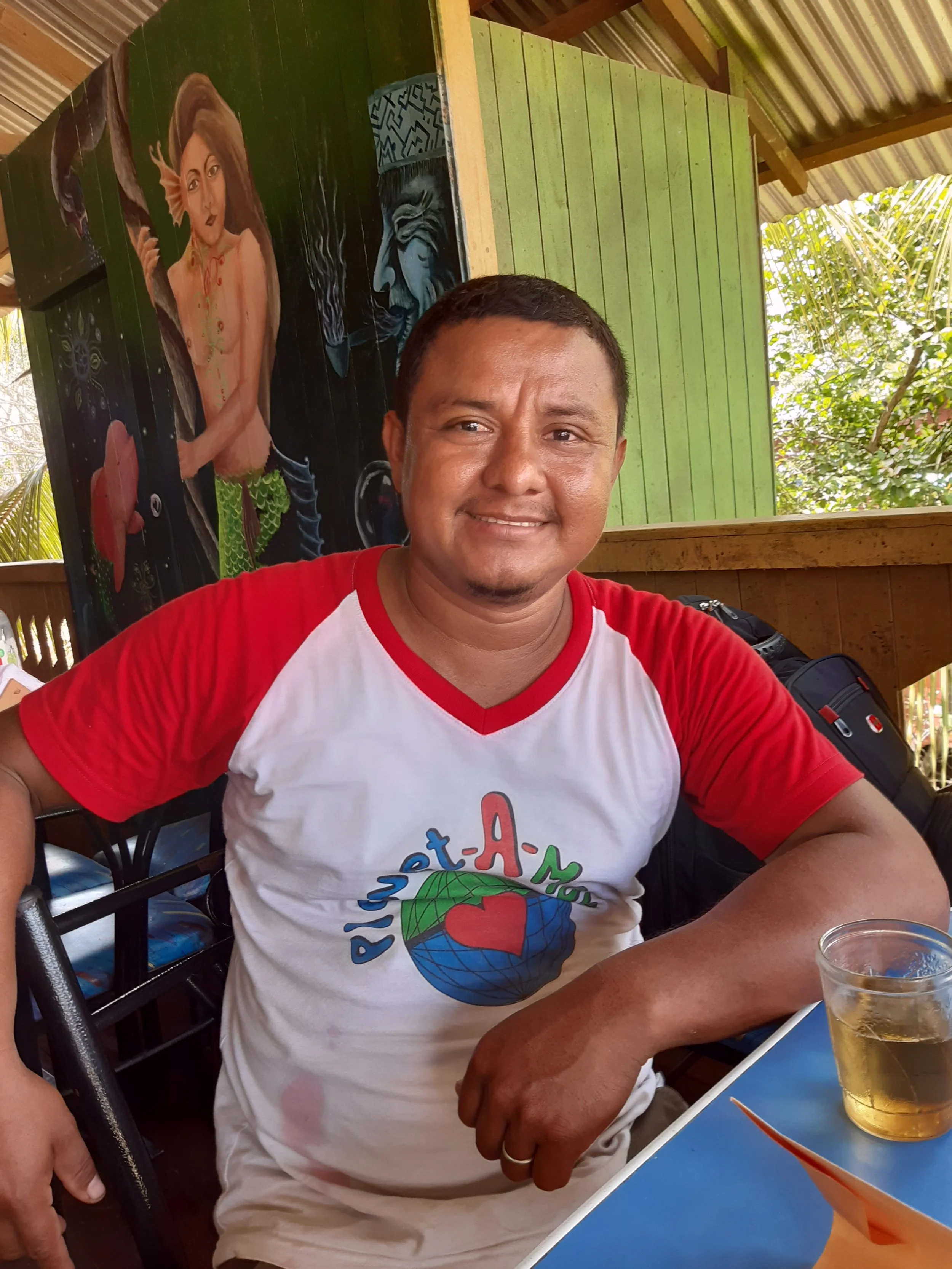 Person wearing a white and red t-shirt with a colorful globe and heart design, seated at a table with a glass of drink. Background includes a mural of a mythical creature on a green wall and tropical plant view.