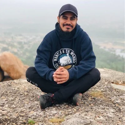 Person sitting cross-legged on a rock wearing a hoodie, cap, and boots, with a blurred landscape in the background.