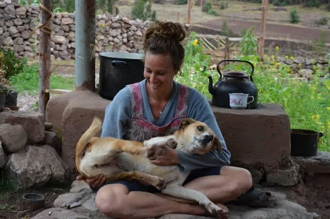 Person sitting outdoors on a stone platform, holding a relaxed dog on their lap, with a teapot and cup nearby.