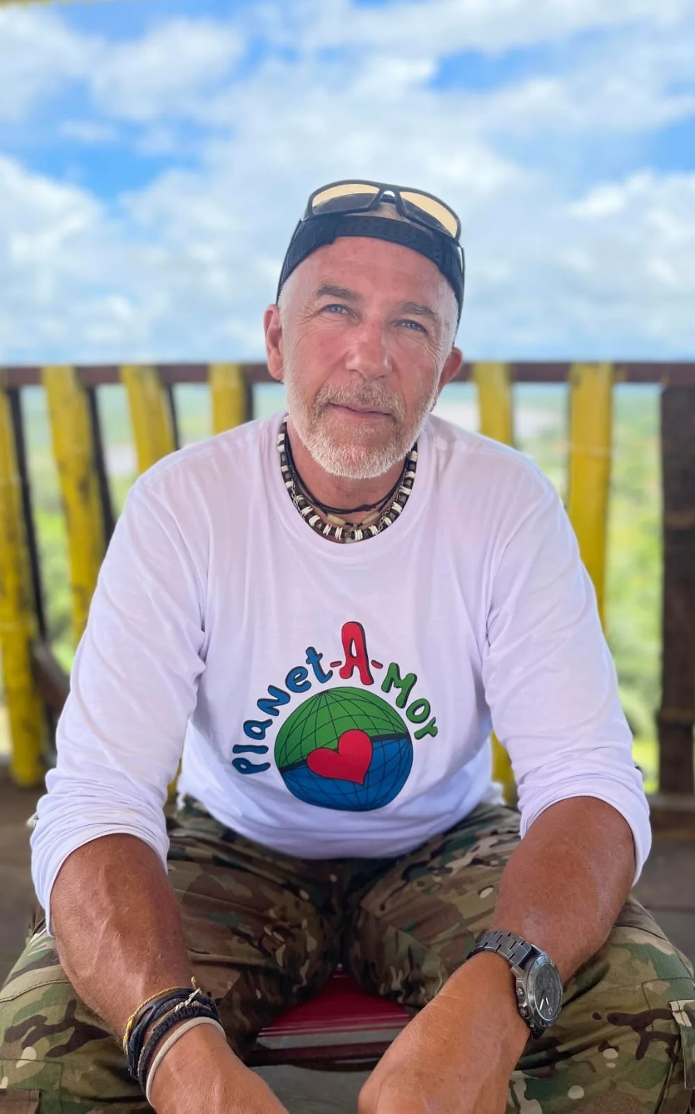 Man sitting wearing a "Planet Amor" shirt, camo pants, beaded necklace, and baseball cap outdoors.