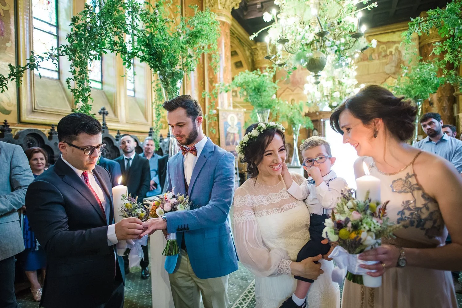 A wedding ceremony taking place in a richly decorated church with gold accents, stained glass windows, and greenery. The bride, smiling and holding a child, is in a white lace dress with a floral crown. The groom, in a light blue suit, is placing a ring on his partner's finger. An officiant in a dark suit stands nearby. Guests observe in the background, some smiling and dressed in formal attire.
