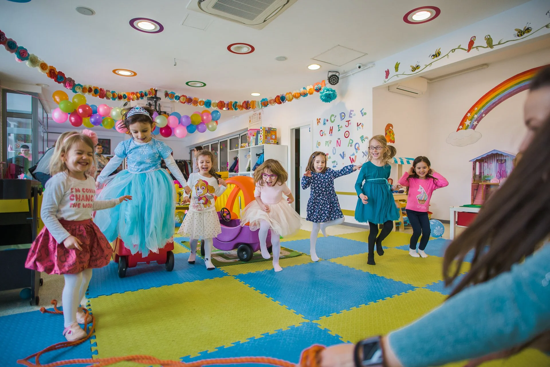 Children at a birthday party playing and jumping in a colorful indoor play area decorated with balloons, streamers, and a rainbow mural.