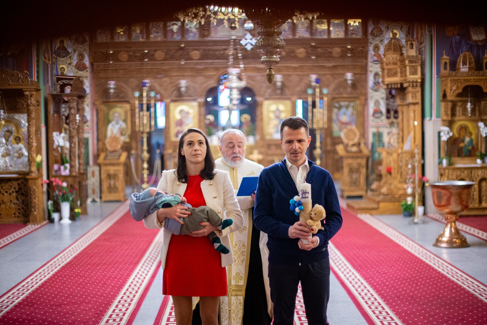 nasul tine lumanarea de botez cu un ursulet pe ea si nasa tine baietulul botezat si de uita la o icoana