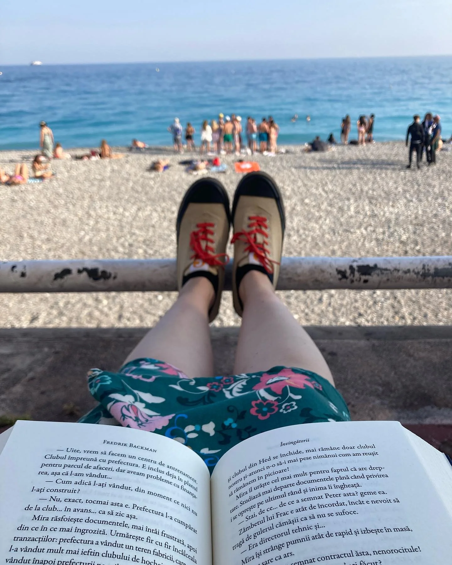 o carte deschisa in prim plan tinuta pe genunchi, cu o plaja si marea in fata