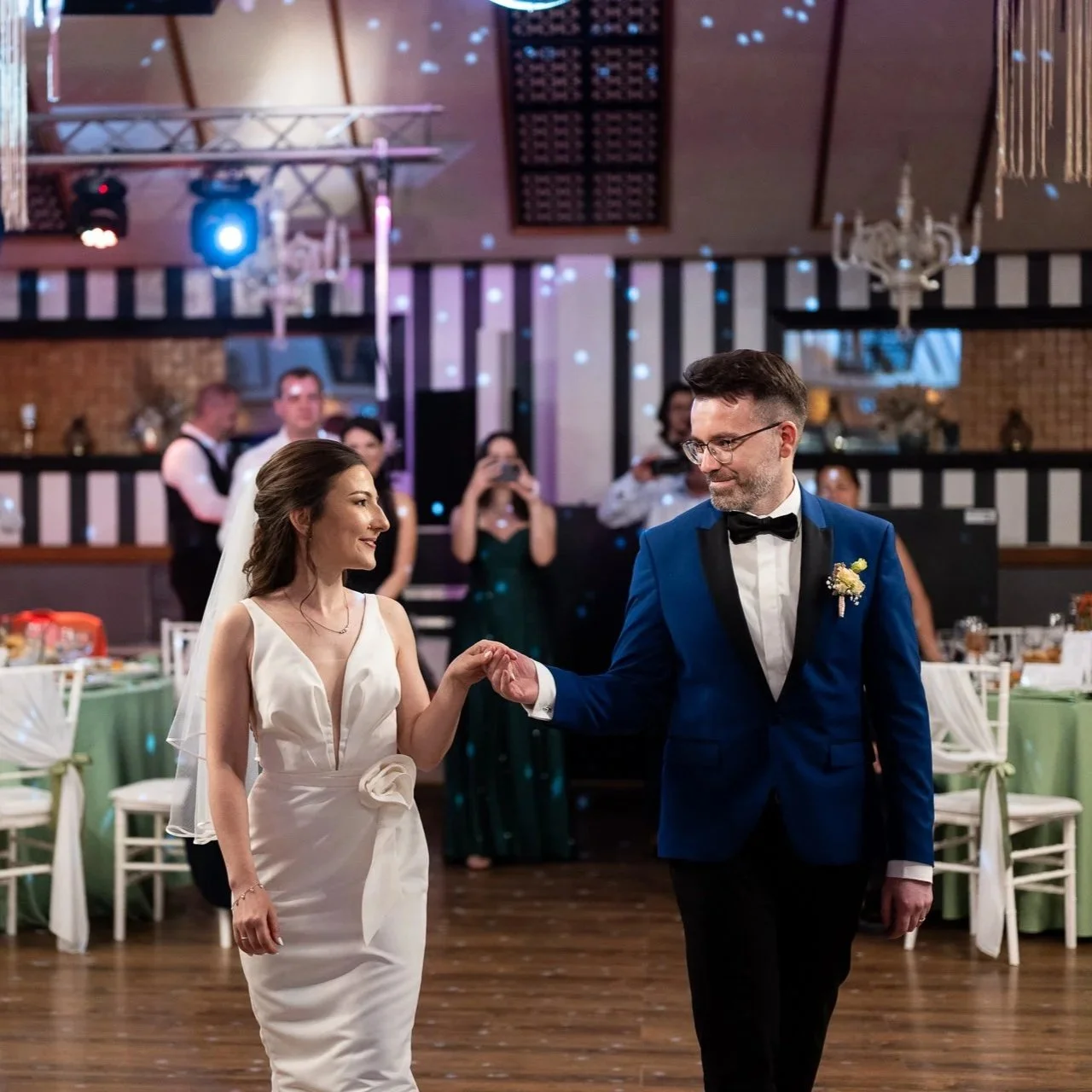 Mirii intră pe ringul de dans la petrecerea de nuntă, ținându-se de mână, sub lumini ambientale și privirile invitaților