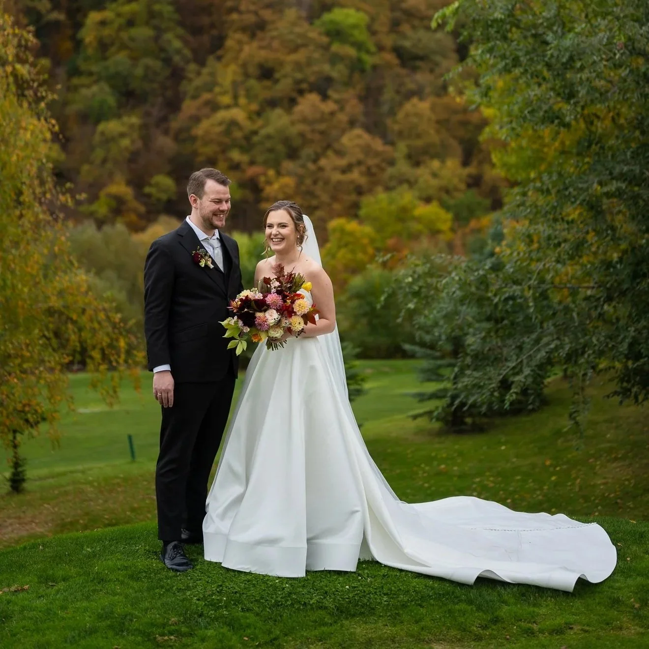 Mireasă și mire stând pe iarbă, într-un peisaj de toamnă, zâmbind, cu pădure colorată în fundal.