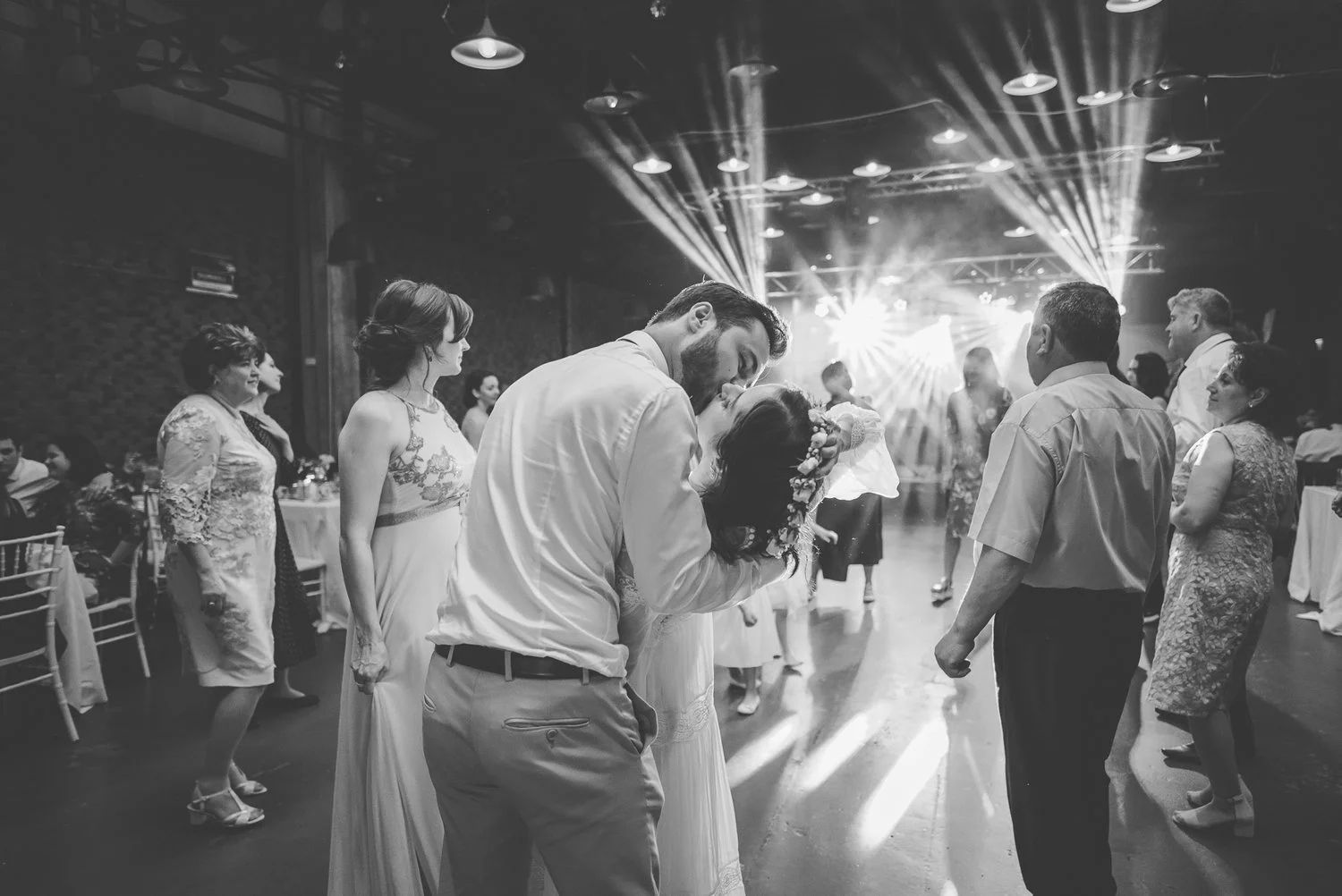 mire si mireasa sarutandu-se pe ringul de dans la petrecerea de nunta inconjurati de invitati iar mireasa isi tine cu mana pe cap o cununa de flori