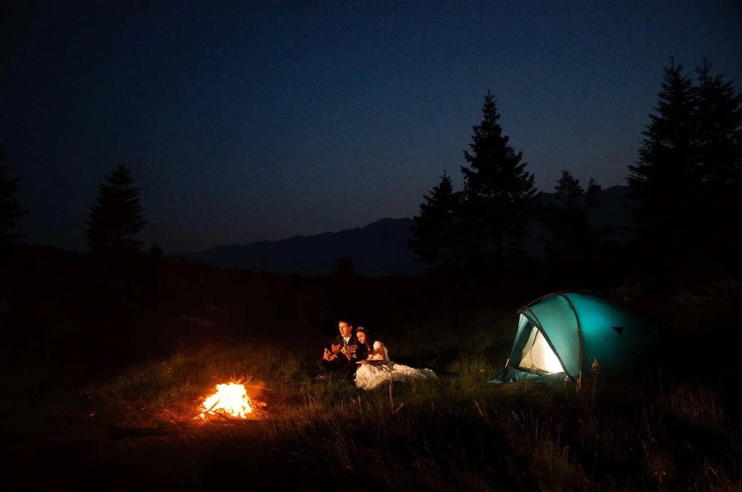 A couple sitting by a campfire at night, with a lit tent nearby and mountains in the background.