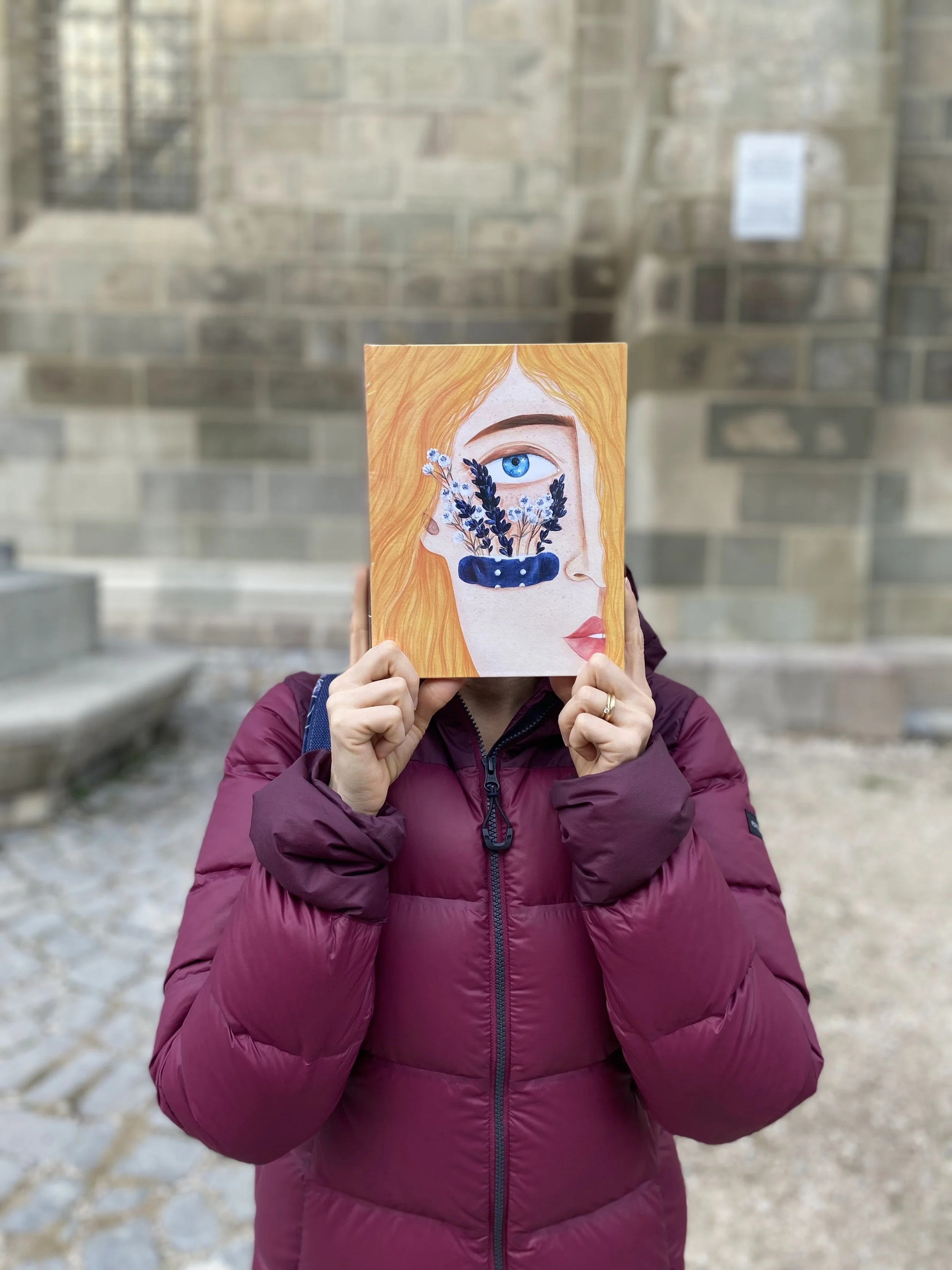 Fata imbracata in geaca de iarna care tine o carte in dreptul feței