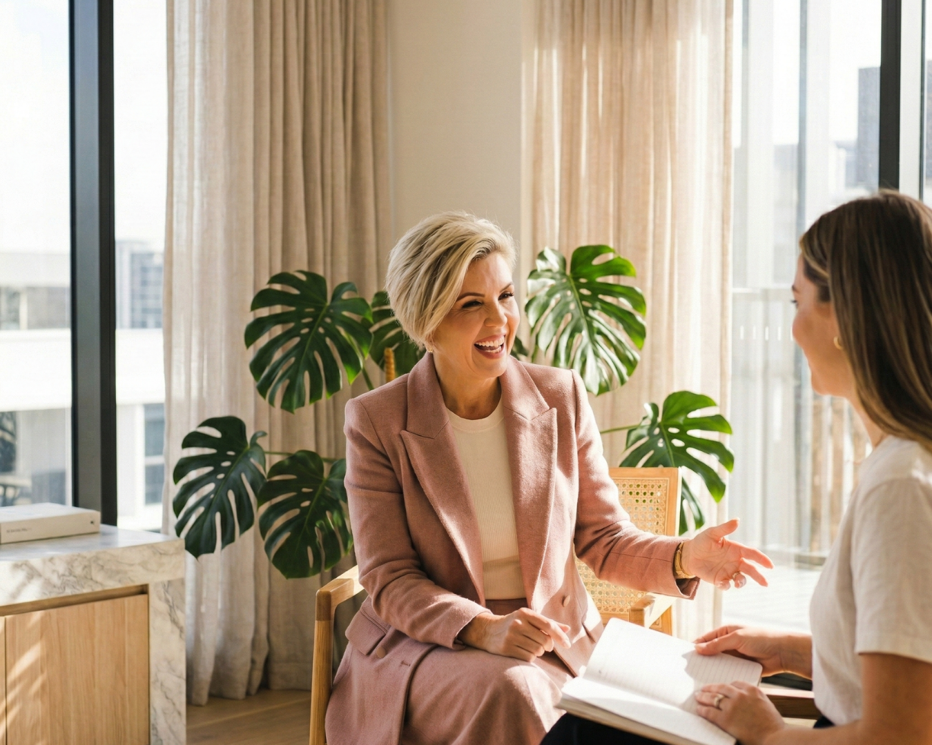 Two women having a conversation in a bright room with large windows, one woman with short blonde hair wearing a pink blazer smiling and gesturing, the other woman with long brown hair holding a notebook. Green Monstera plants behind them.