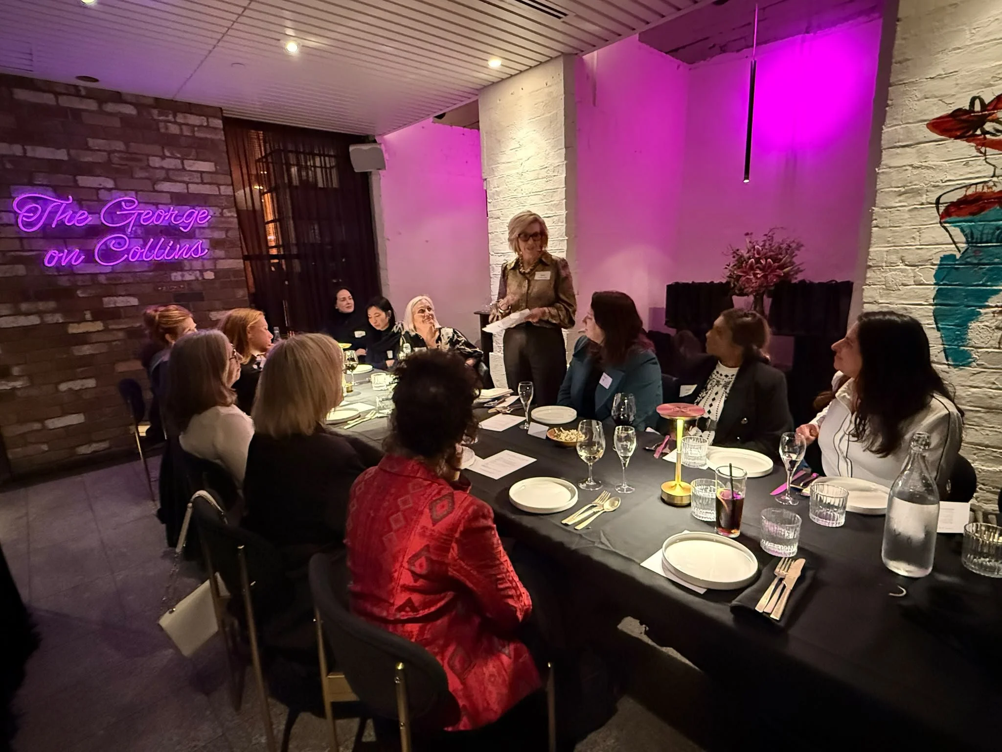 A group of women sitting at a long banquet table in a restaurant or event space, listening to a woman standing and speaking. The setting has exposed brick and white painted walls, with pink and purple decorative lighting and a neon sign that reads 'The George on Collins.' There are wine glasses, plates, and utensils on the table.