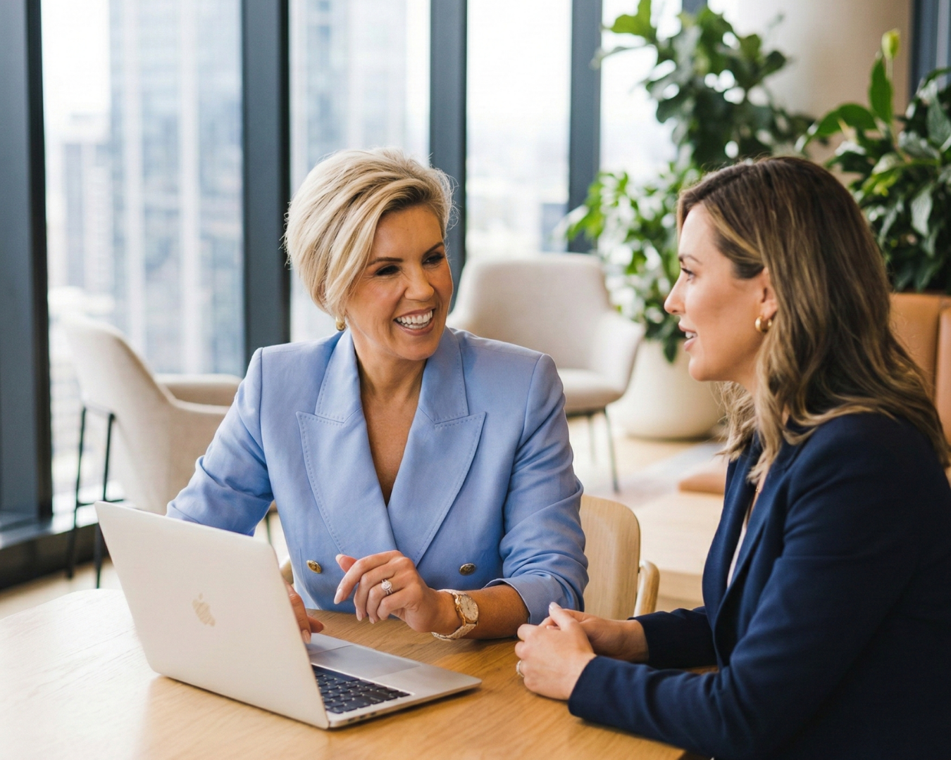 Two women having a conversation at a table in a bright office space with large windows and green plants. One woman is wearing a light blue blazer, and the other is wearing a dark blazer. There is a silver laptop open on the table.