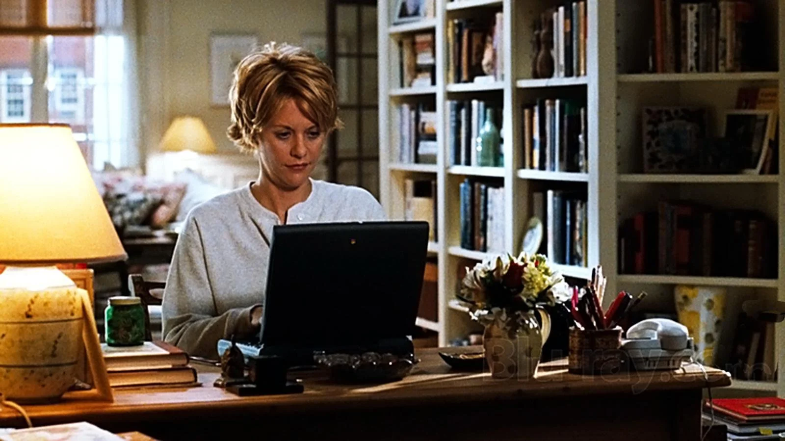 A woman with short blonde hair working on a laptop at a wooden desk in a cozy, well-lit home office or living space that has bookshelves filled with books and decorative items, as well as a bed and lamps in the background.