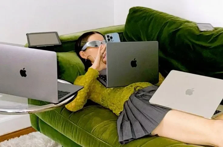 A woman lying on a green couch surrounded by multiple Apple laptops and a tablet, wearing virtual reality glasses while holding one of the devices.