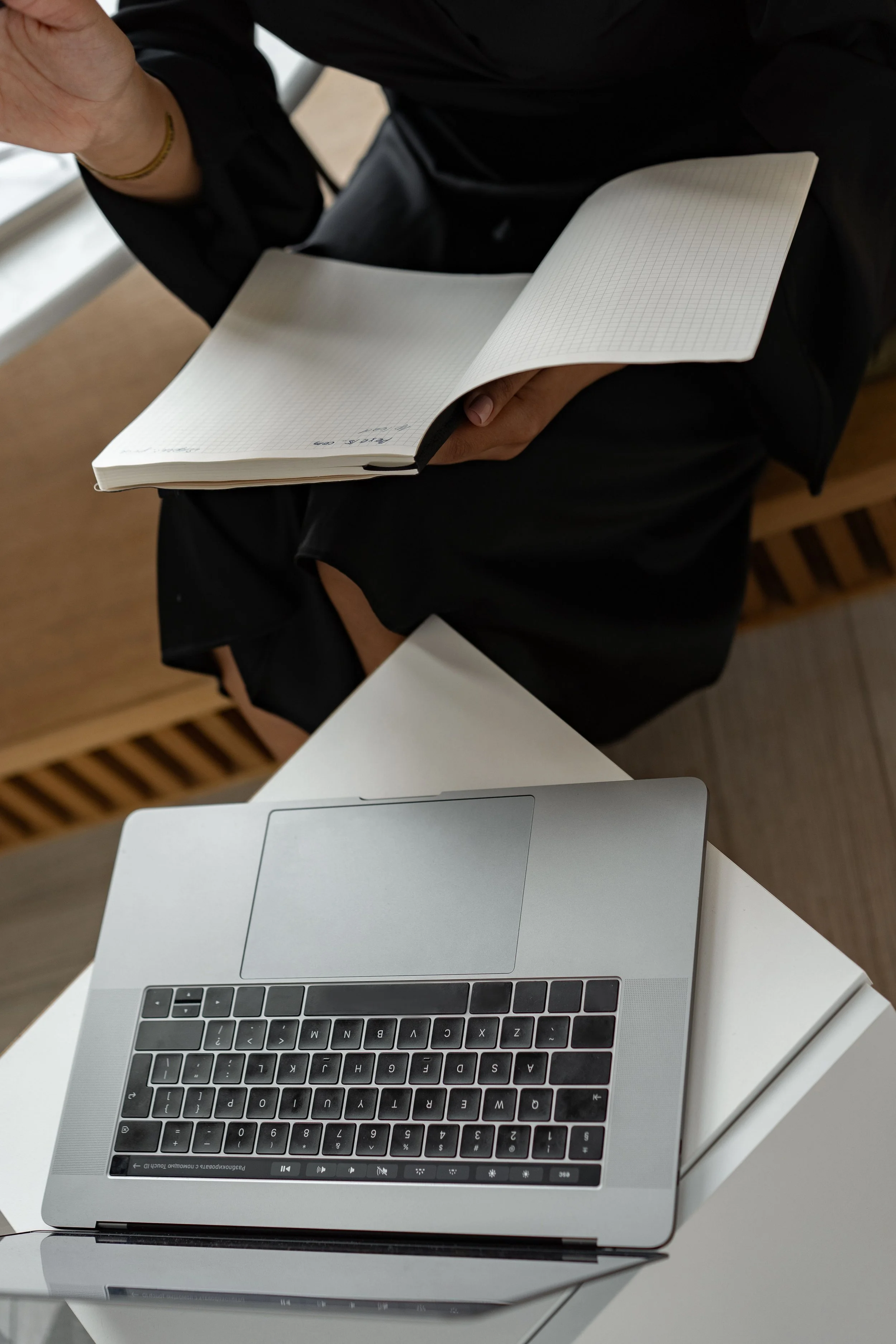 A person in black clothing holding an open notebook with grid paper, sitting at a white desk with a silver laptop in front, on a brown wooden floor near a window.