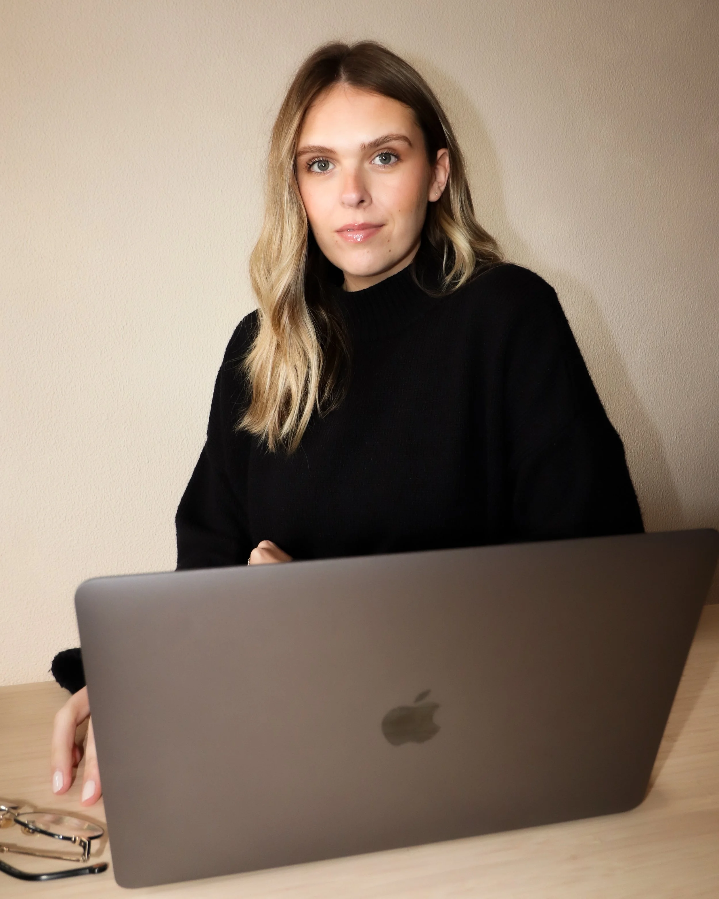 A young woman with long blonde hair and blue eyes sitting at a desk with a closed laptop in front of her, holding a pair of glasses in her left hand, against a plain beige wall background.