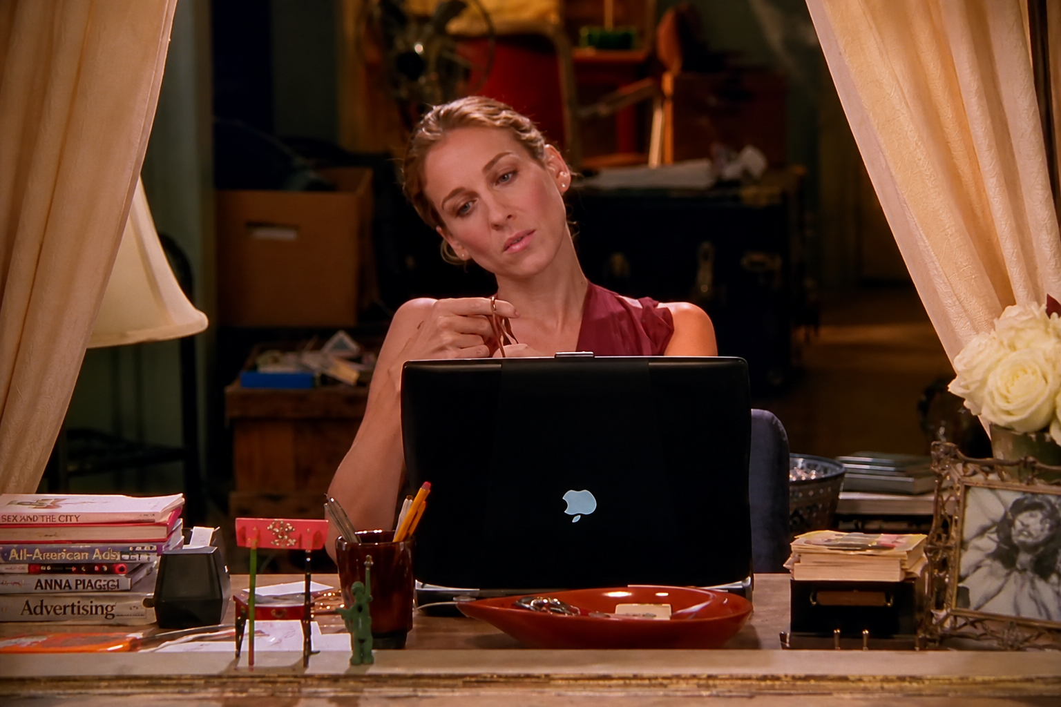 A woman with blonde hair, wearing a sleeveless maroon top, sits at a cluttered desk looking thoughtfully at her open laptop. The desk has books, a small sculpture, a container with pencils, a tray with rings, and a framed photo of a woman in black and white. Two light-colored curtains frame the scene.