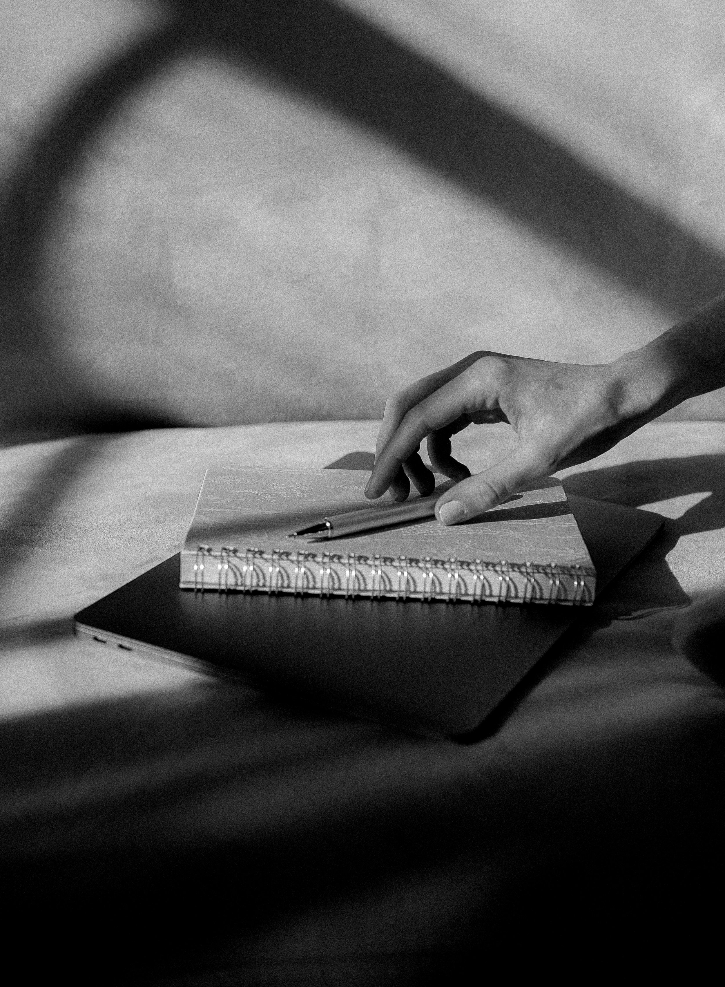 Close-up of a hand reaching to pick up a pen from a notebook on a desk, with a spiral-bound notebook and papers nearby in black and white.