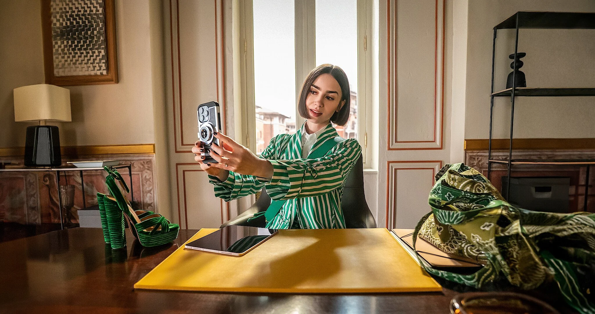 A woman with shoulder-length dark hair and a striped green and white blazer sitting at a desk, looking at her phone, in a room with patterned walls and a large window behind her.