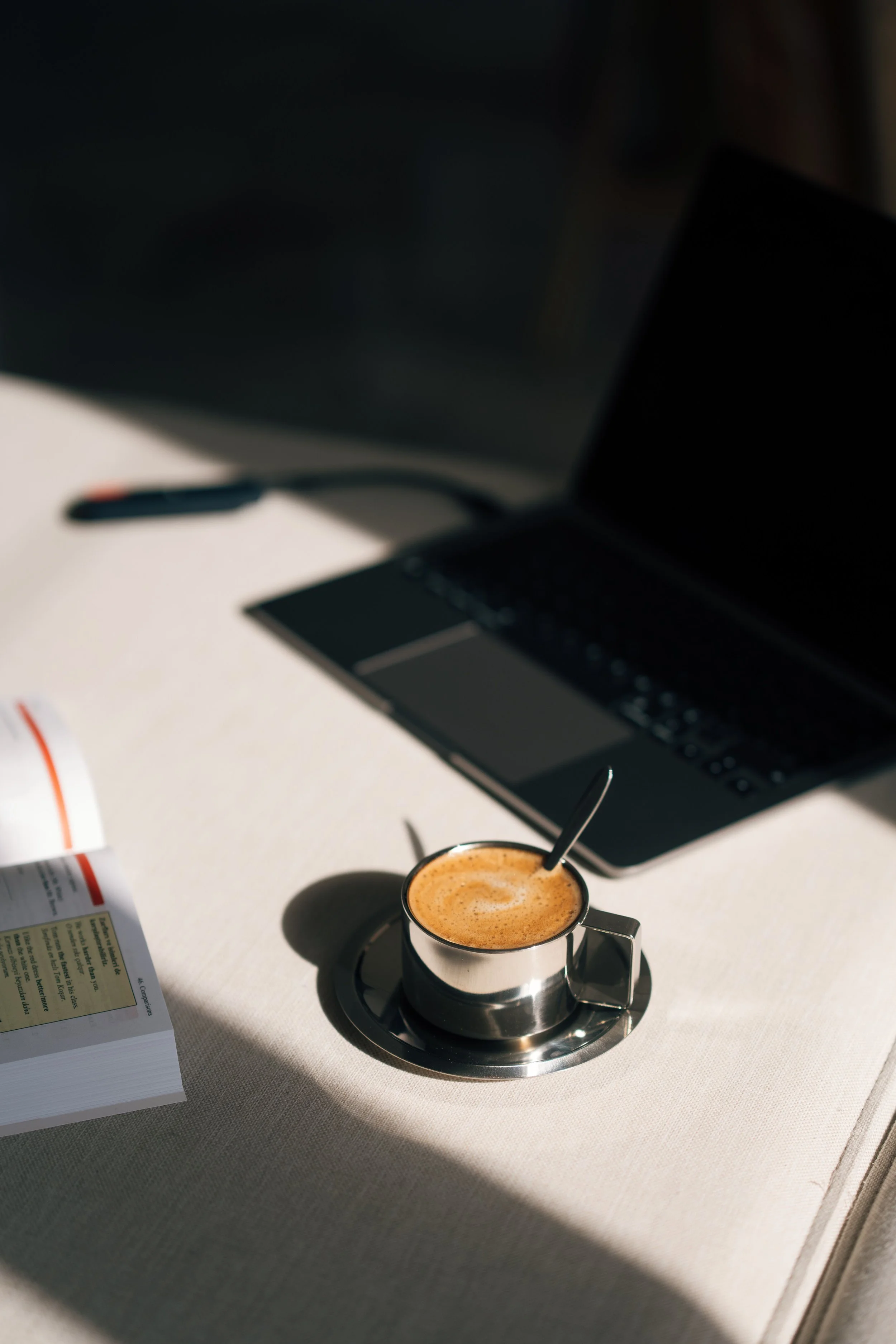 A cup of coffee with foam on top placed on a metal saucer on a light-colored table, with a laptop in the background and some papers and a pen nearby.