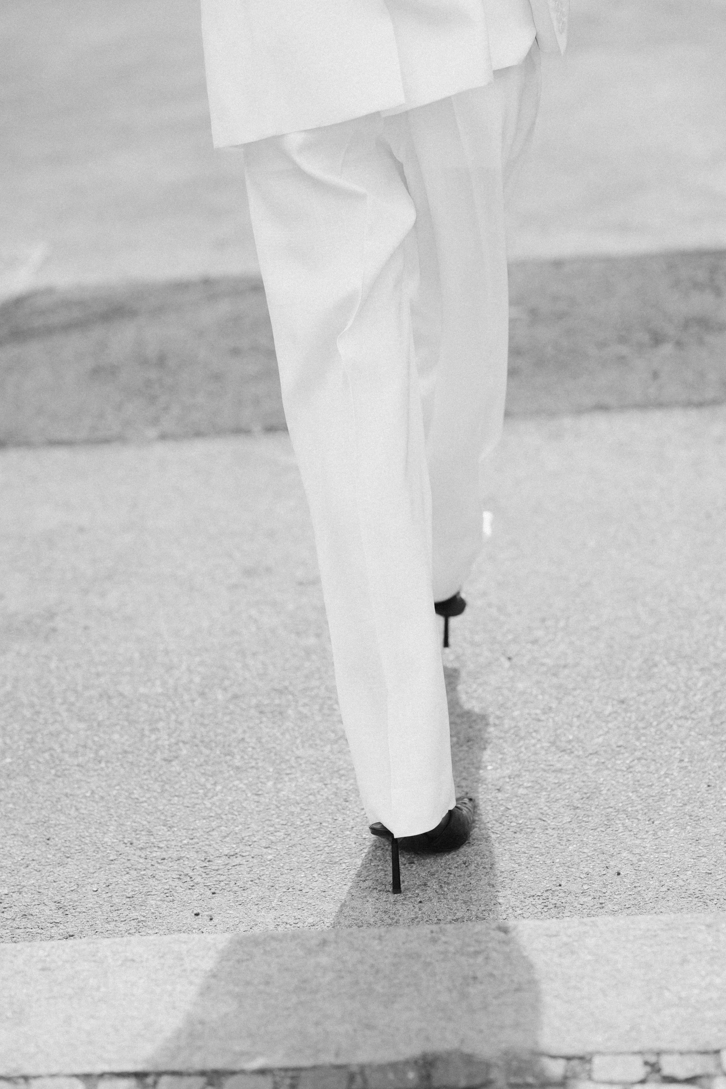 A person wearing long white pants and black shoes, holding a cane, standing on a textured walkway.