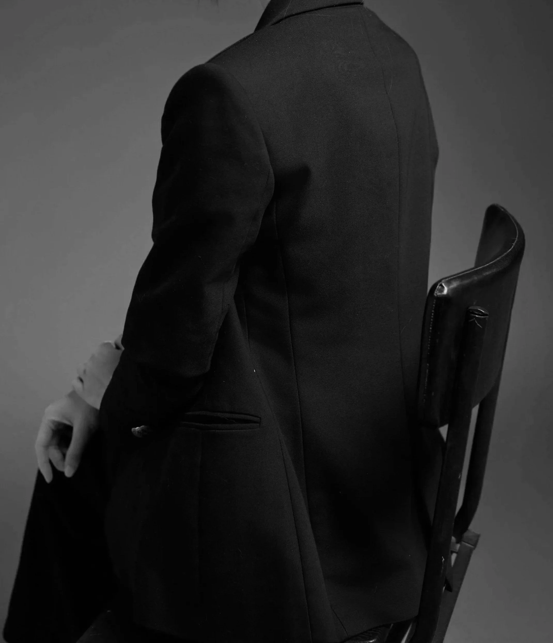 Black-and-white image showing a person in a suit sitting on a chair, facing sideways with their face not visible. The person's right arm is resting on their lap, and part of the arm and leg are visible.