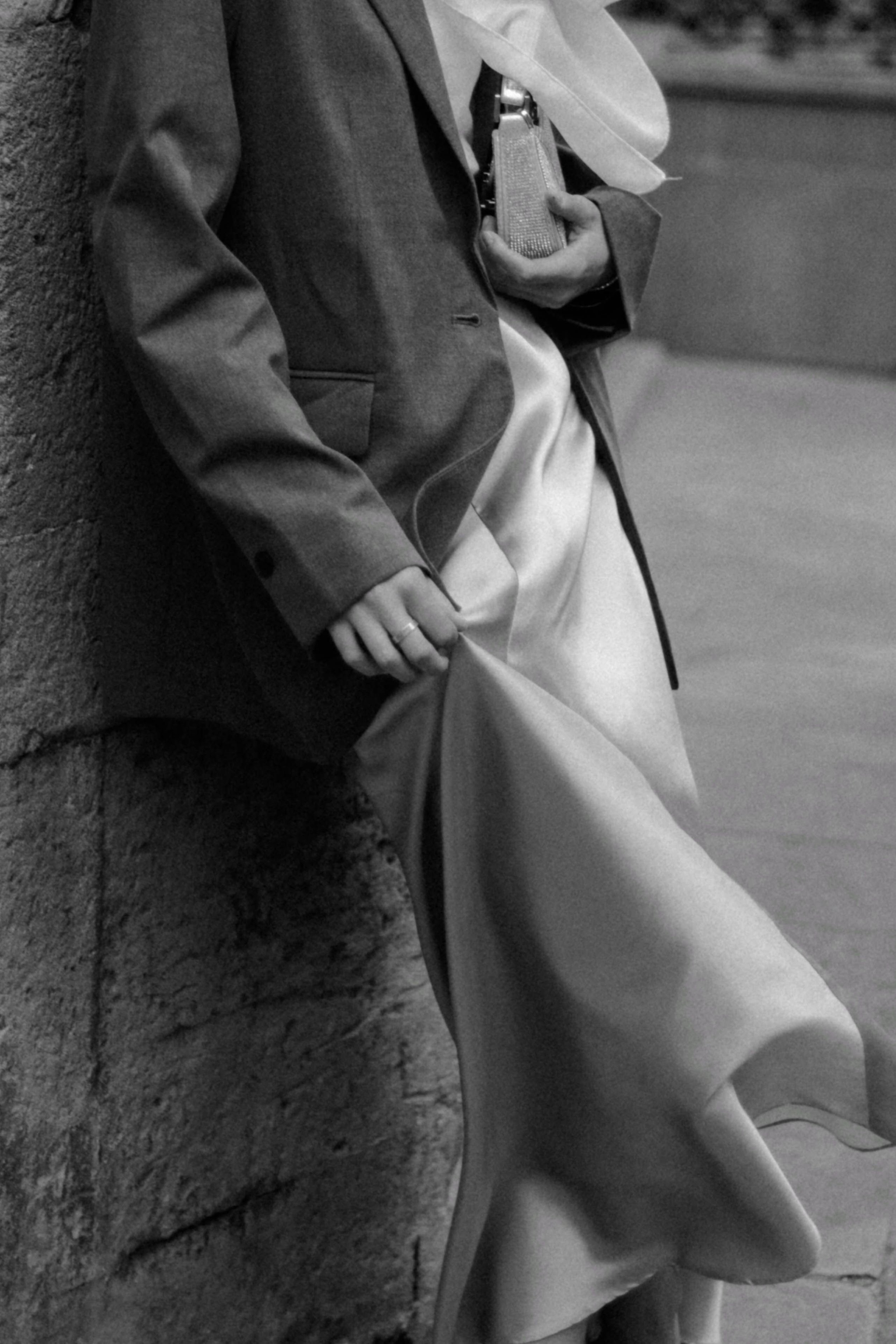 Close-up of a person leaning against a textured wall, holding a small clutch purse, wearing a blazer and light-colored pants, with part of the face visible and the background blurred.