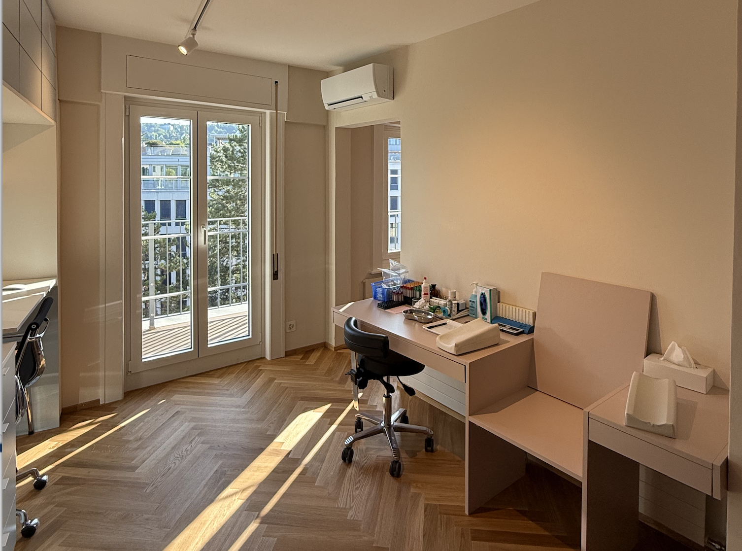 Labor der Hausarztpraxis mit einem Schreibtisch, einem Stuhl, einer Fensterfront und Parkettboden.