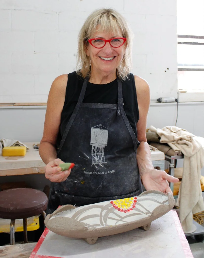 Artist Suze lindsay smiles at the camera from the waist up, wearing red glasses and an apron, a piece of in progress handmade pottery in front of her on the studio table.