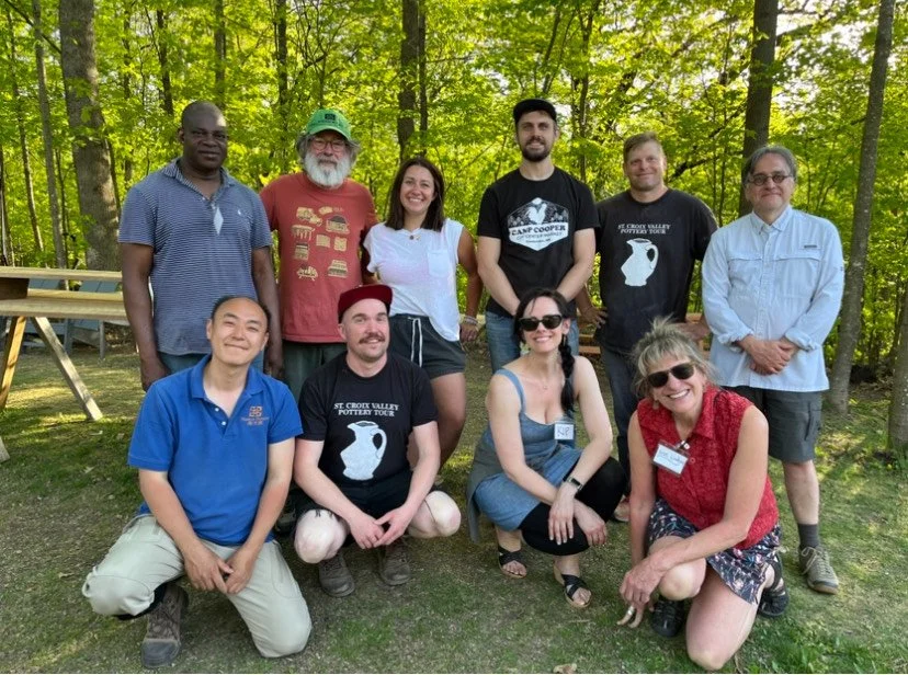 A group of potters in two rows, some crouching and some standing against a green summer-tree background.
