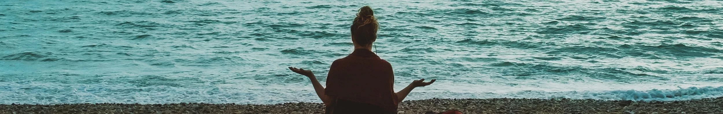 Person sitting cross-legged on a pebble beach with arms outstretched, facing the ocean.