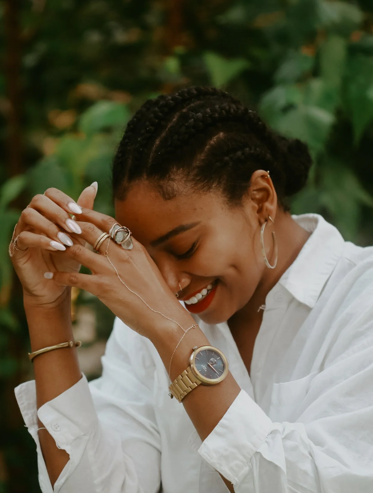 A woman with braided hair smiling, wearing a white shirt, gold watch, and silver jewelry, with her hands clasped near her face, set against a blurred green foliage background.