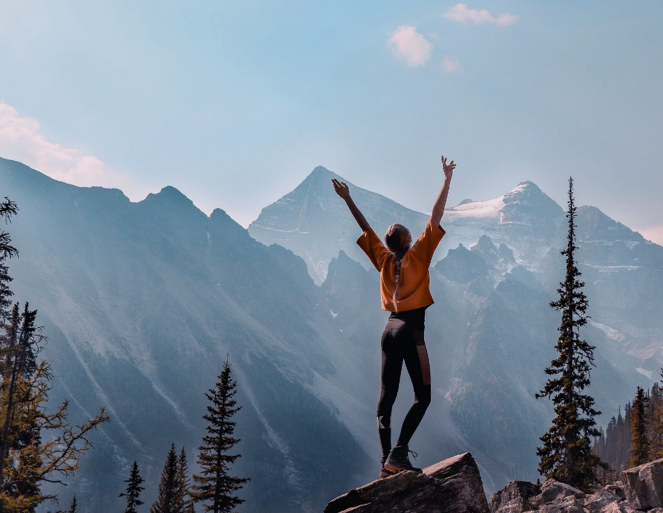 A person standing on a rock with arms raised, overlooking a mountain range with snow-capped peaks and tall pine trees.