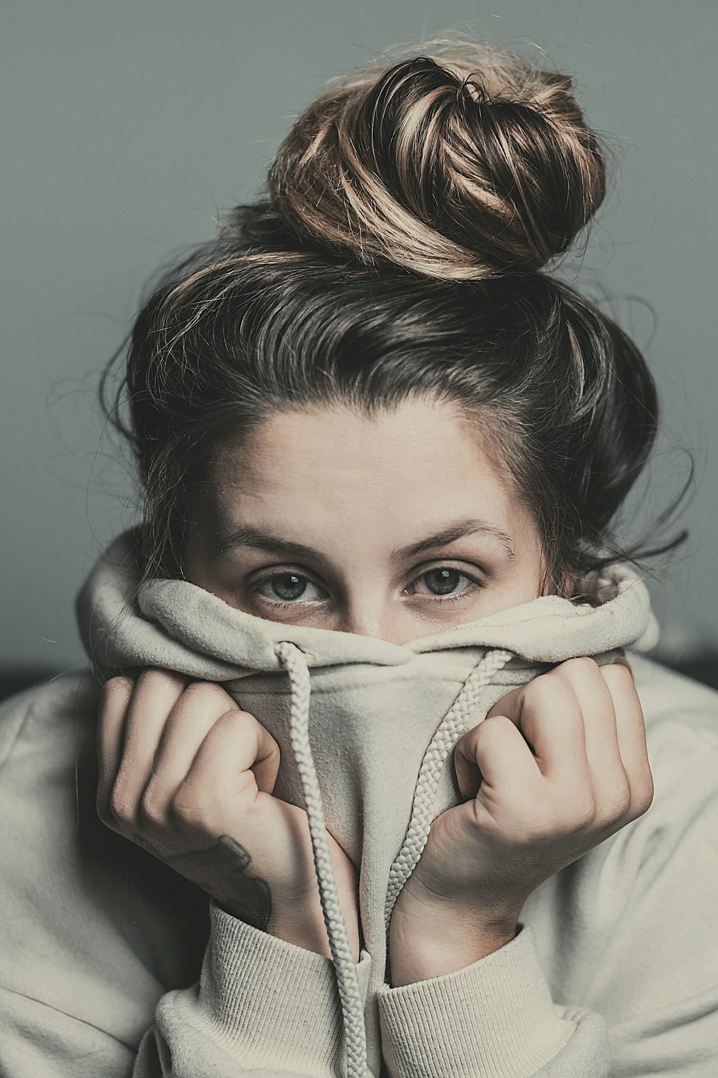 A woman with blue eyes and light skin, partially hiding her face with a beige hoodie, is looking directly at the camera. Her hair is styled in a messy bun on top of her head, with highlights and dark brown shades. She is holding the hoodie with both hands, pulling it up around her face.