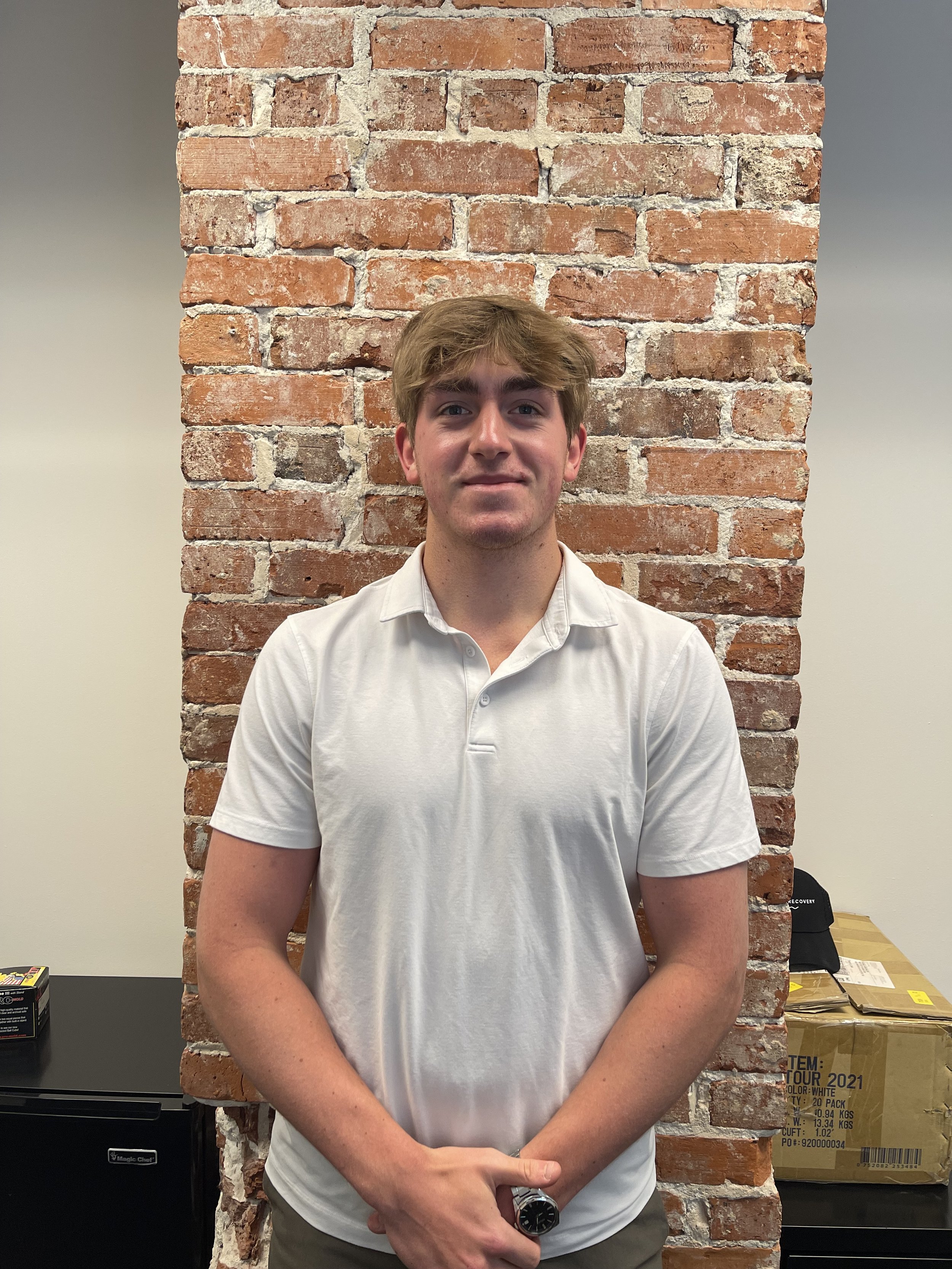 A man with wavy brown hair smiling at the camera, wearing a white top, with a brick stone wall in the background.