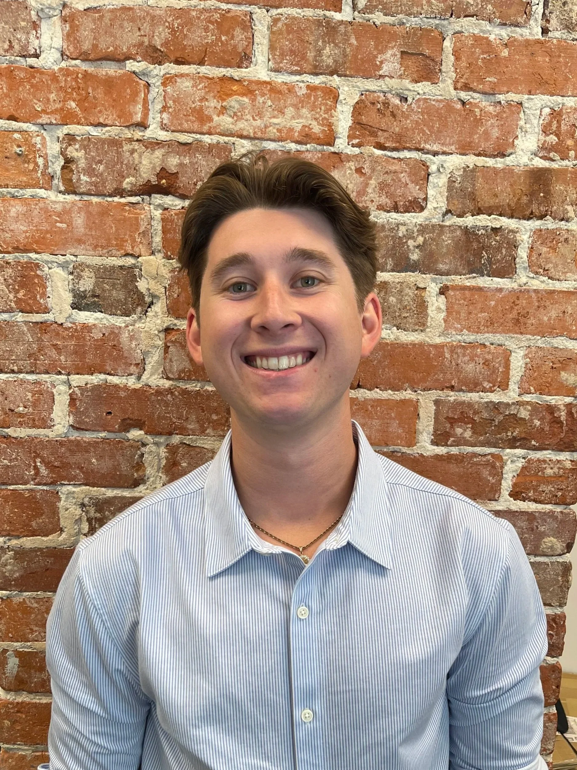 A young man with short dark hair and light skin, wearing a blue shirt with a sailboat pattern and a gold chain, standing in front of a brick wall.