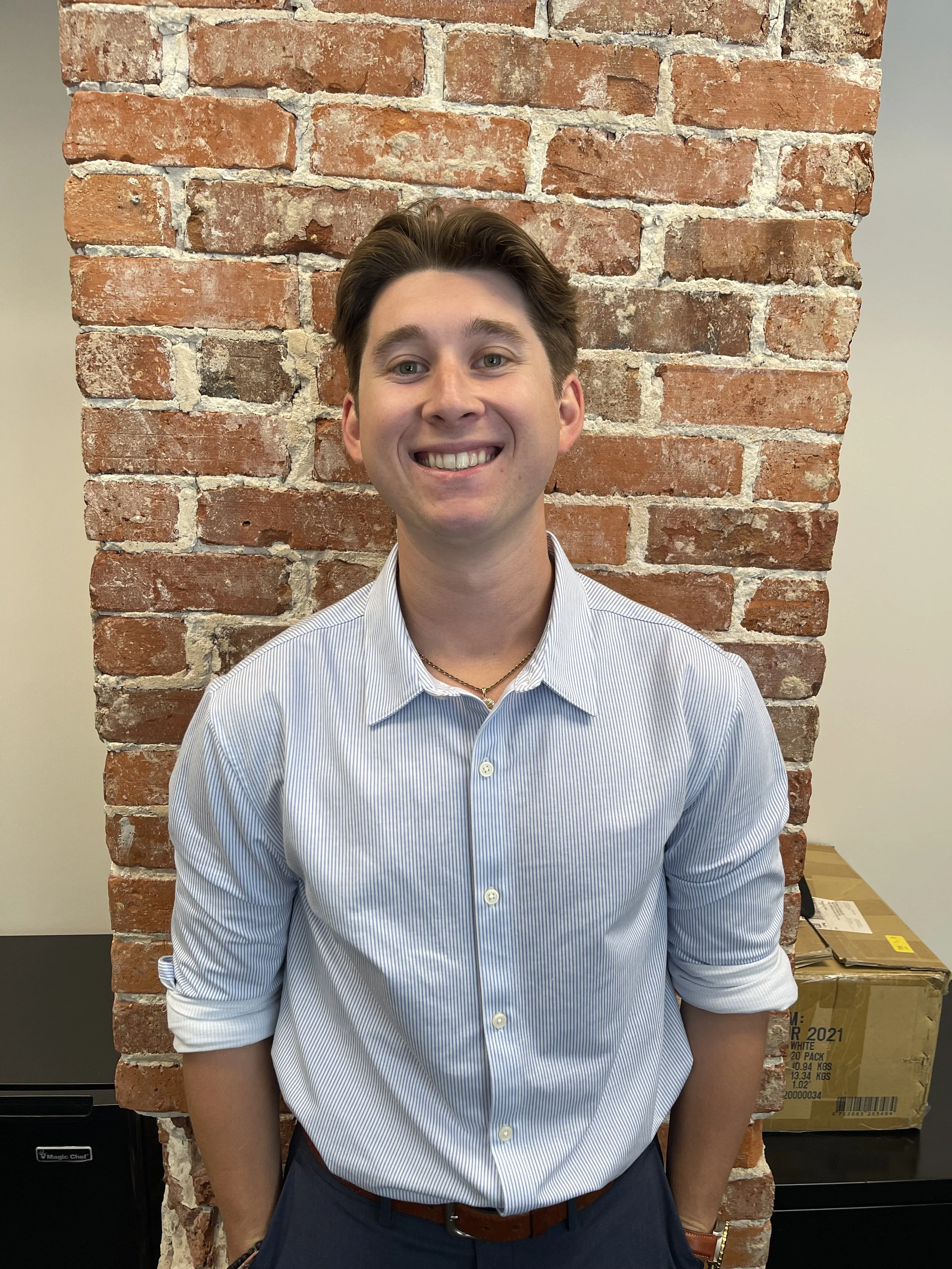 A young man with short dark hair and light skin, wearing a blue shirt with a sailboat pattern and a gold chain, standing in front of a brick wall.