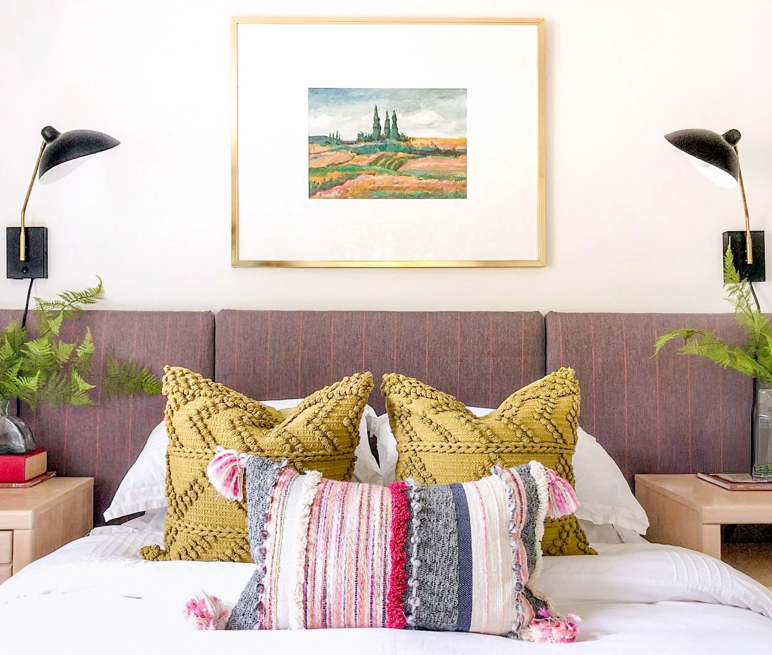 A bedroom with two bedside tables, each with a houseplant, black wall-mounted reading lamps, a purple upholstered headboard, colorful pillows, and a framed landscape painting on the wall.