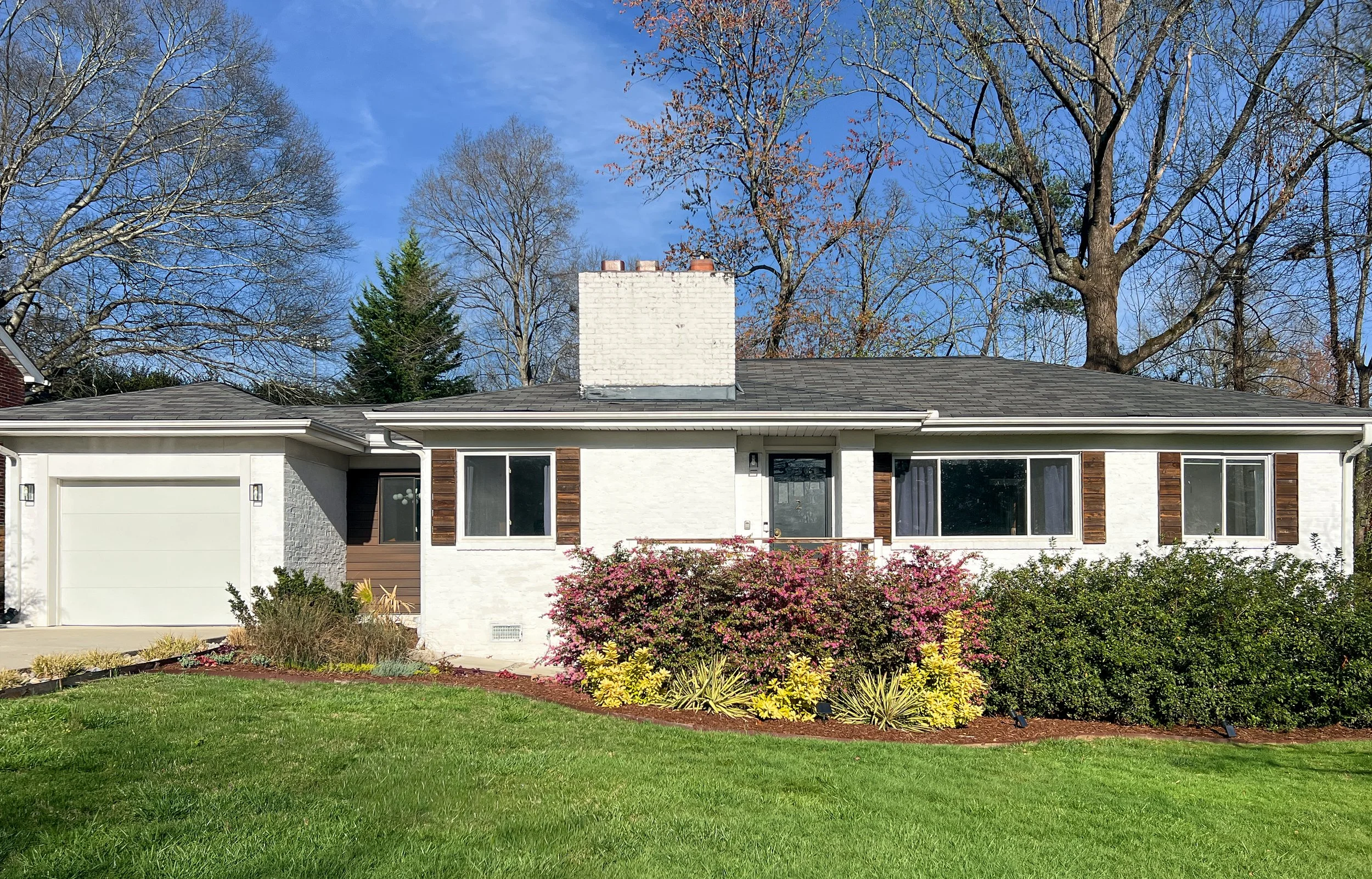 Front view of a single-story white house with dark roof, large windows with wooden shutters, a well-kept garden with colorful bushes, and a green lawn, under a blue sky with some trees in the background.