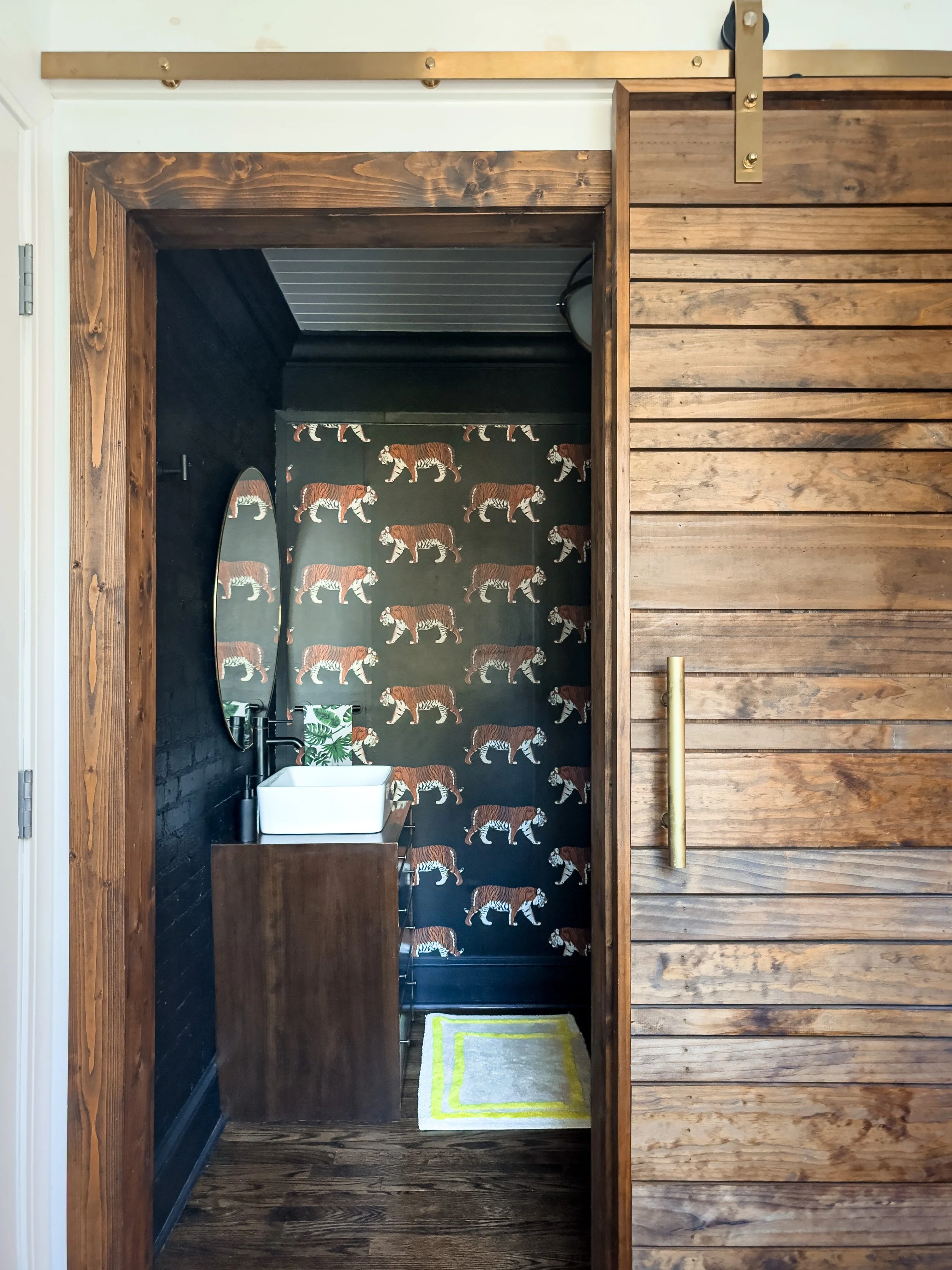 Small bathroom with dark walls featuring a tiger wallpaper accent wall, a white square vessel sink, a tall wooden vanity, a mirror, a black soap dispenser, a green and white decorative item, a small rug with yellow and white borders, and a closet door with wood panels.