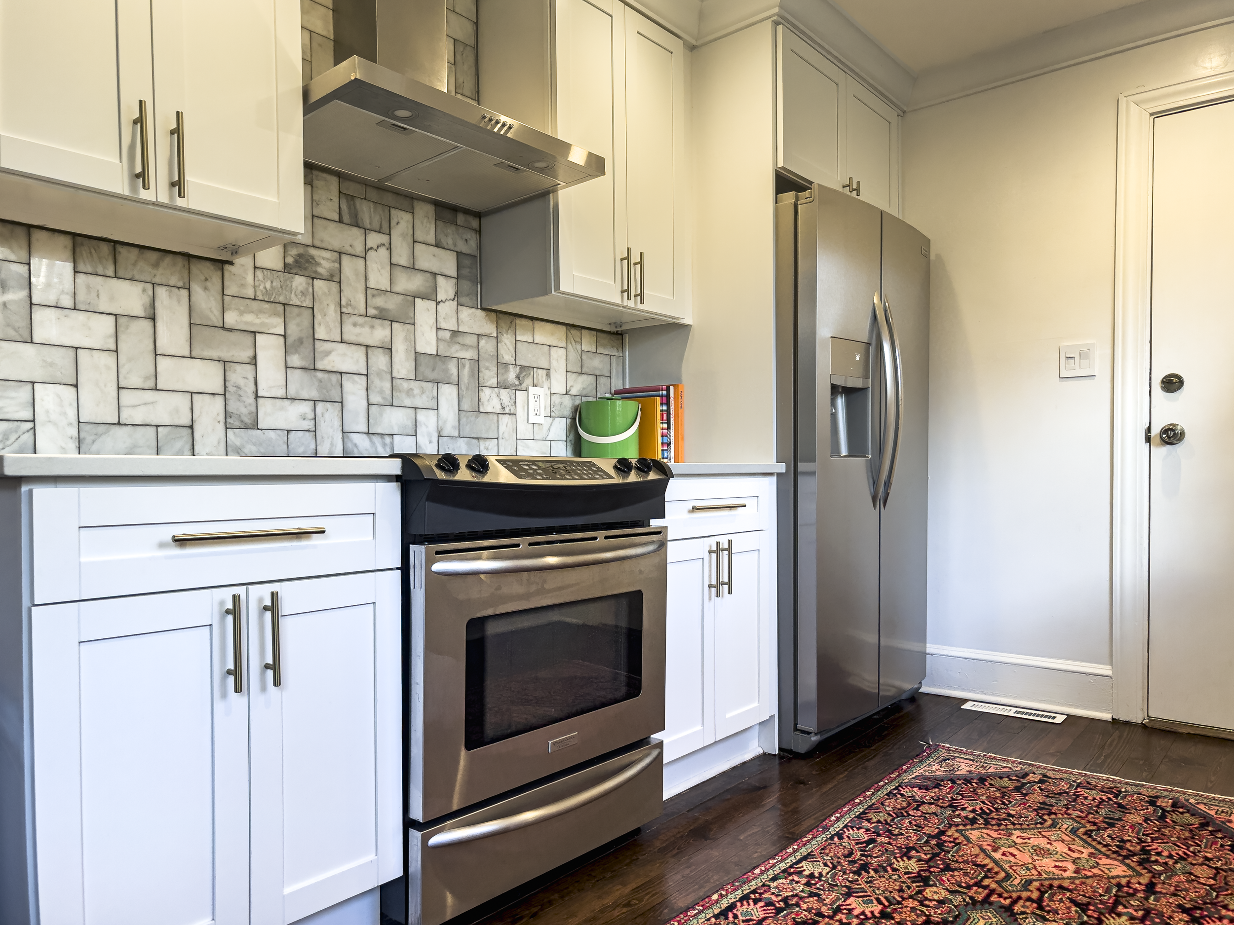 Kitchen with stainless steel refrigerator and stove, white cabinets, tile backsplash, colorful books and a green container on the countertop, dark wood floor, patterned rug, white door with doorknob and lock.