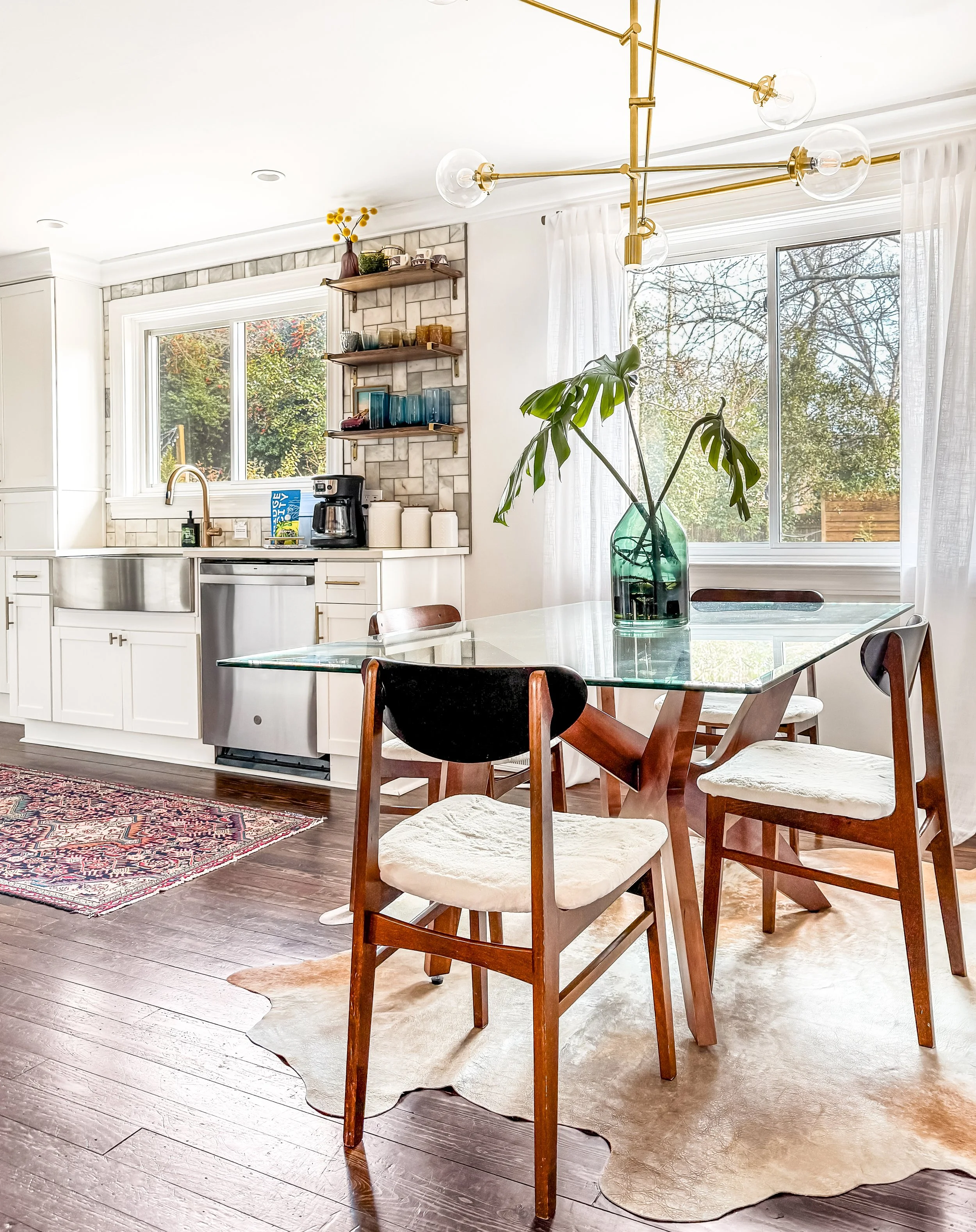 A bright, modern kitchen and dining area with a glass-top table, four wooden chairs with white cushioned seats, a large window with white curtains, a green plant in a glass vase, open wooden shelves with cups and glasses, and a patterned rug on the hardwood floor.