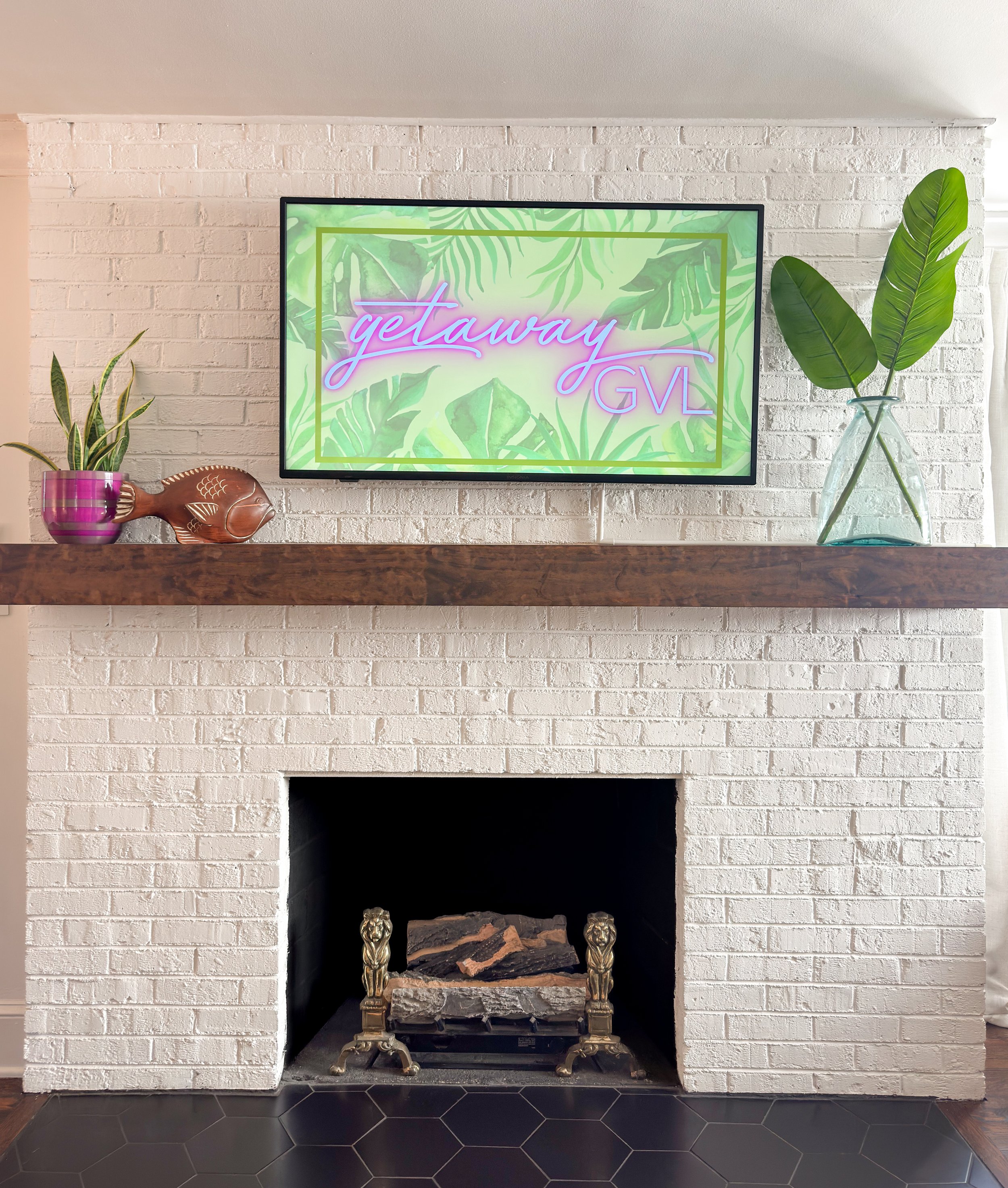 Living room fireplace with a wooden mantel, decorated with plants and a fish-shaped sculpture, a TV above displaying a tropical leaf background with the words "getawayGVL" in pink and purple neon style.