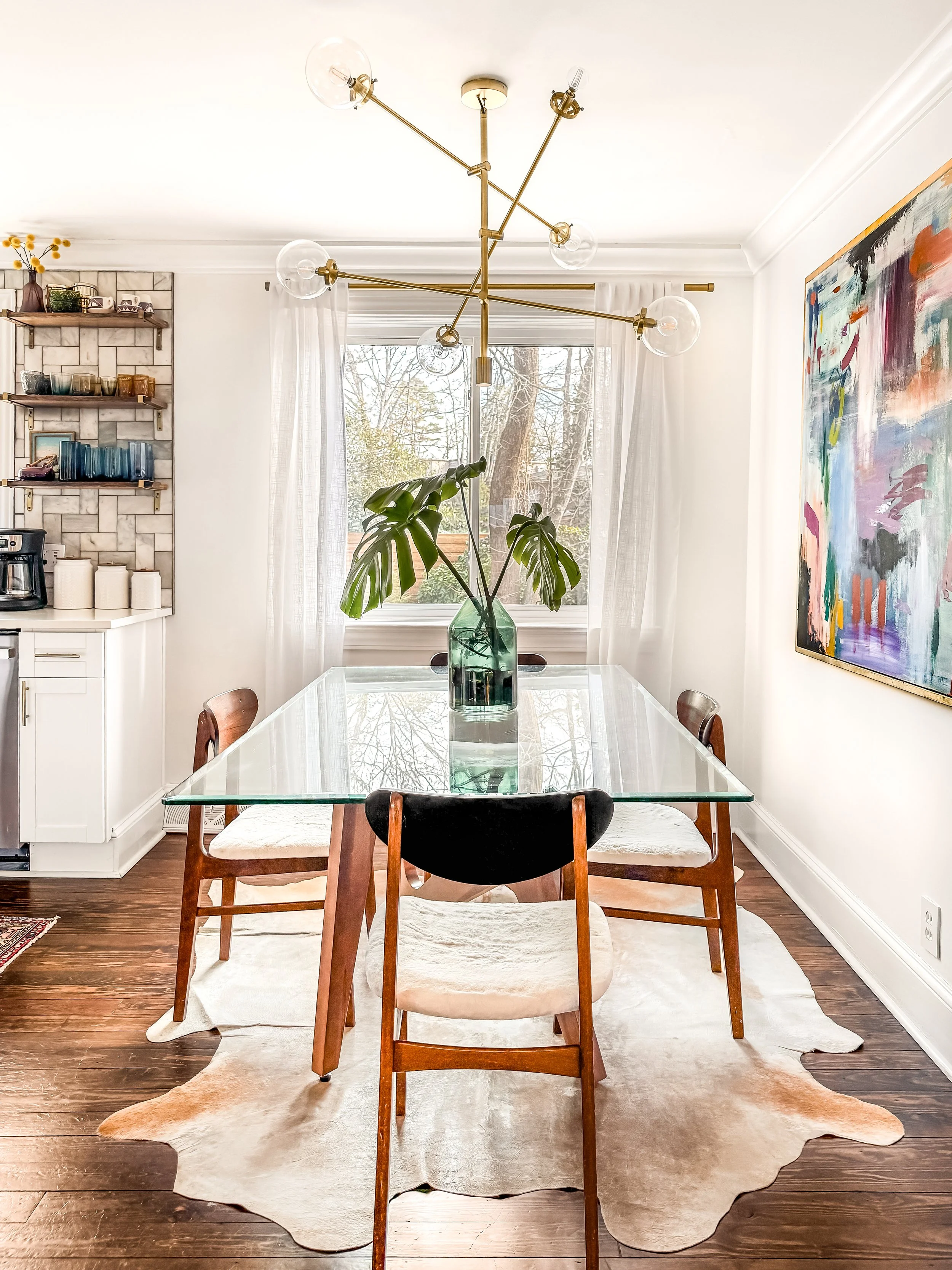 A dining room with a glass-top table, four chairs, a large green vase with Monstera leaves, a modern gold chandelier, a window with white curtains, an abstract painting on the wall, a shelf with dishes, and a dark wood floor.